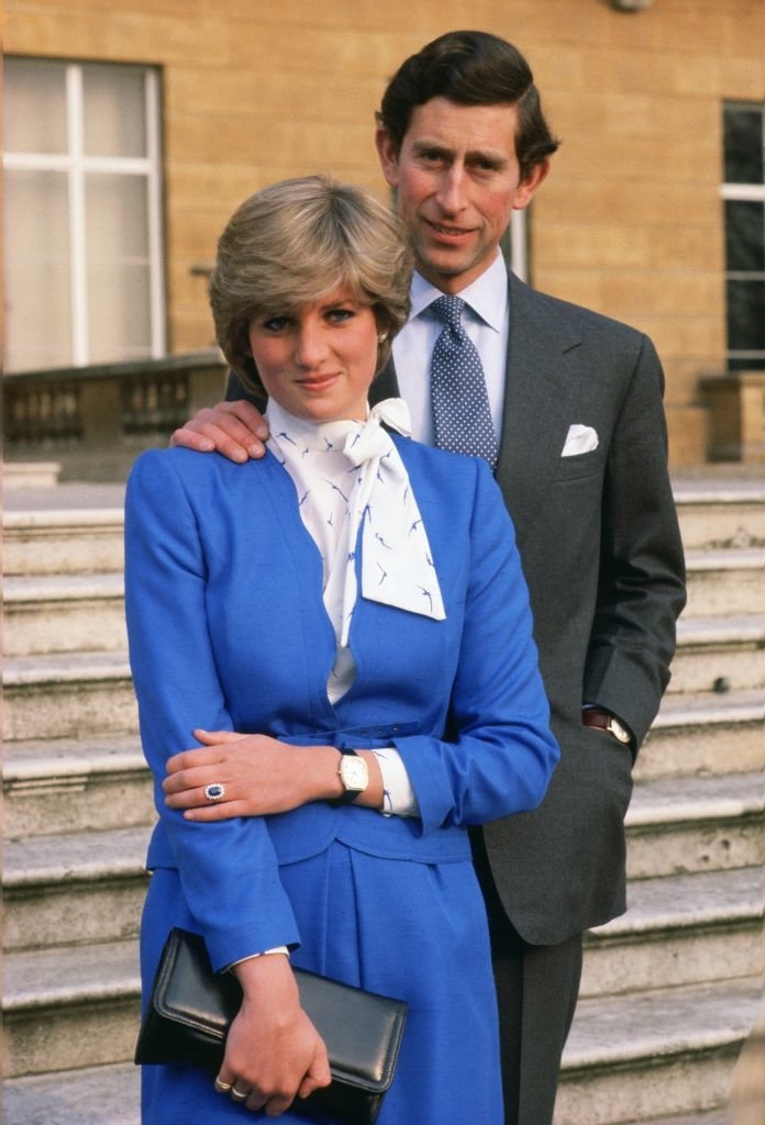 Princess Diana in a blue outfit and white scarf stands with Prince Charles in a grey suit. He has one hand on her shoulder. They stand in front of steps