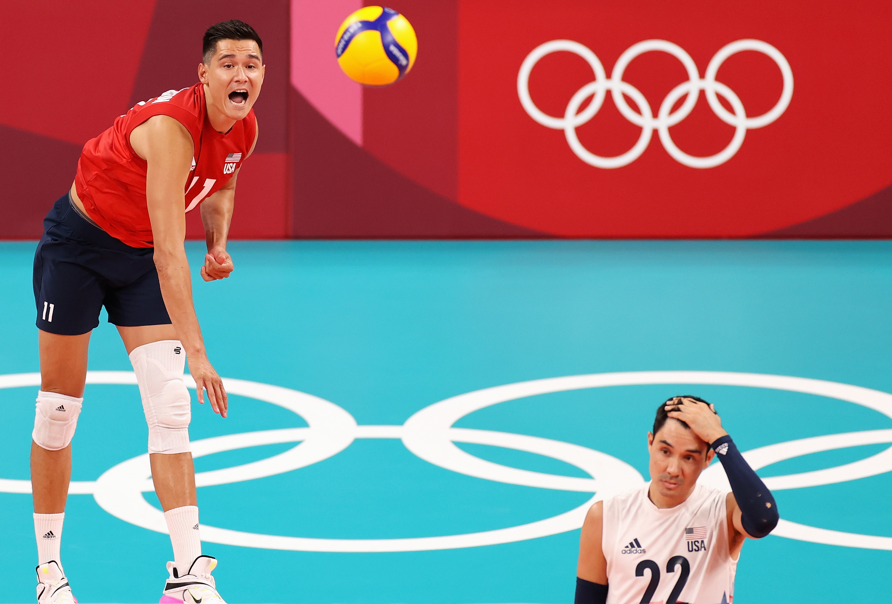USA volleyball players with the number 11 jumping to spike a ball and number 22 crouching on the court during an Olympic match