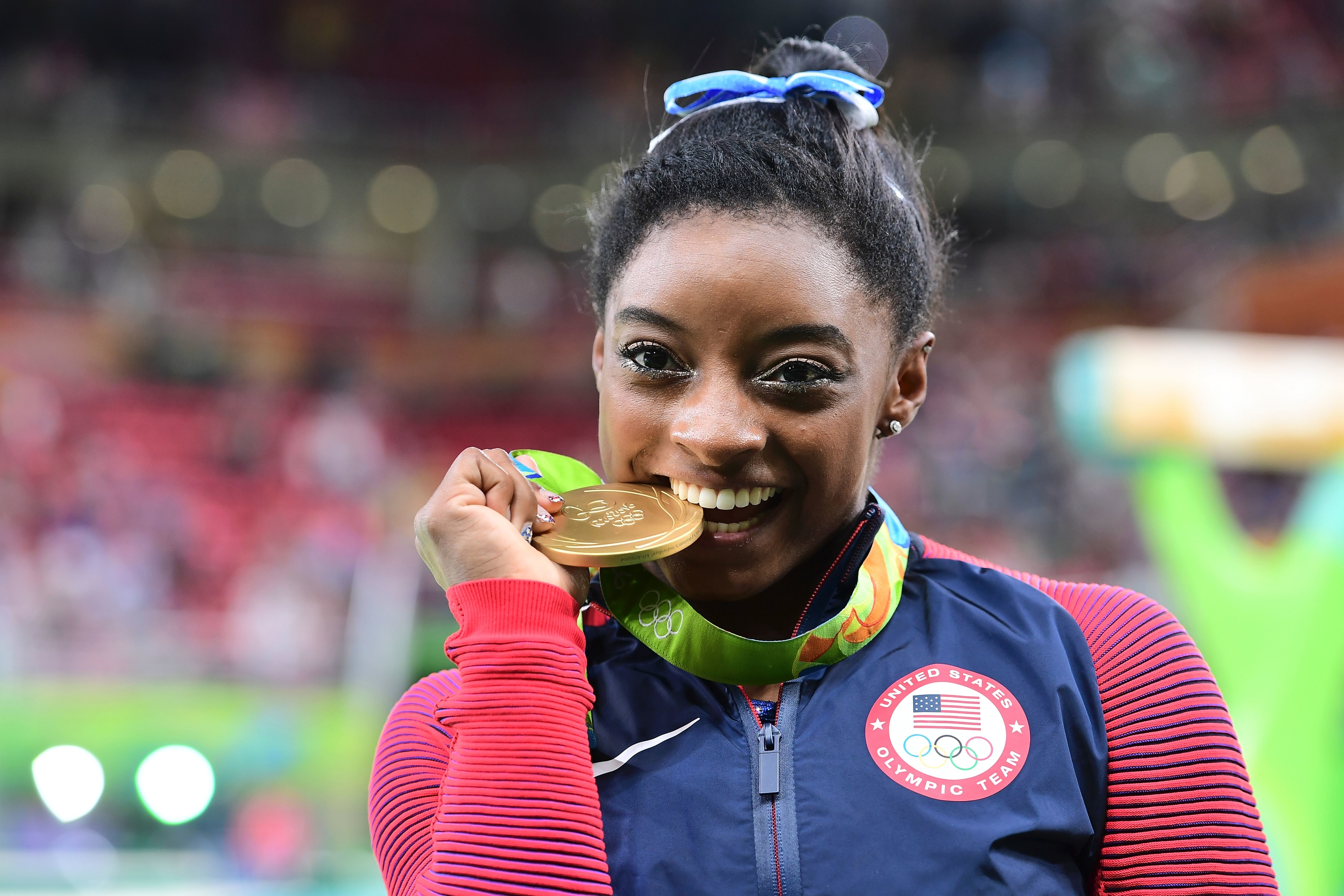 Simone Biles, wearing Team USA gear, bites her gold medal at the Olympics, smiling with joy