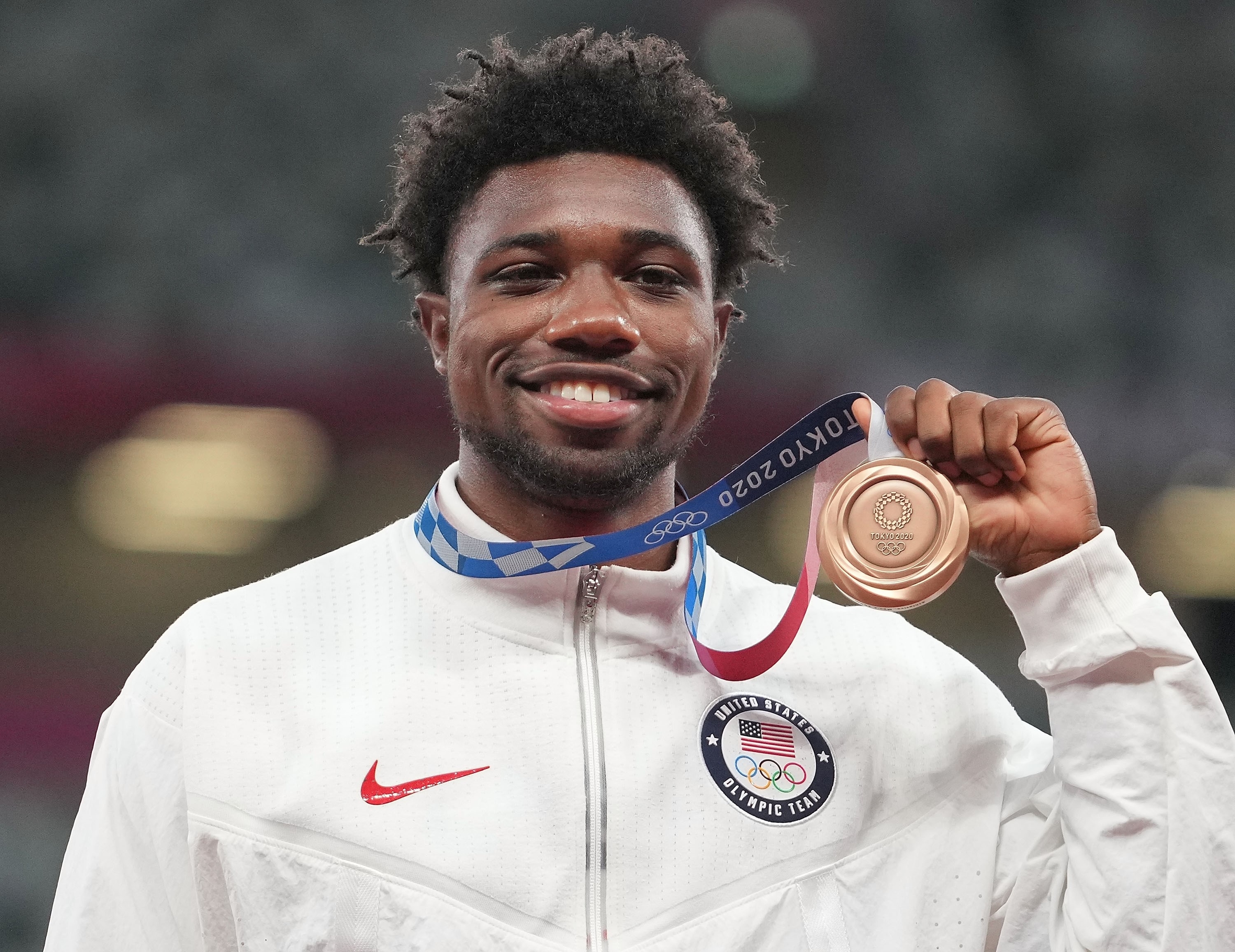 Noah Lyles holds up a Tokyo 2020 bronze medal, wearing a white USA Olympic Team jacket