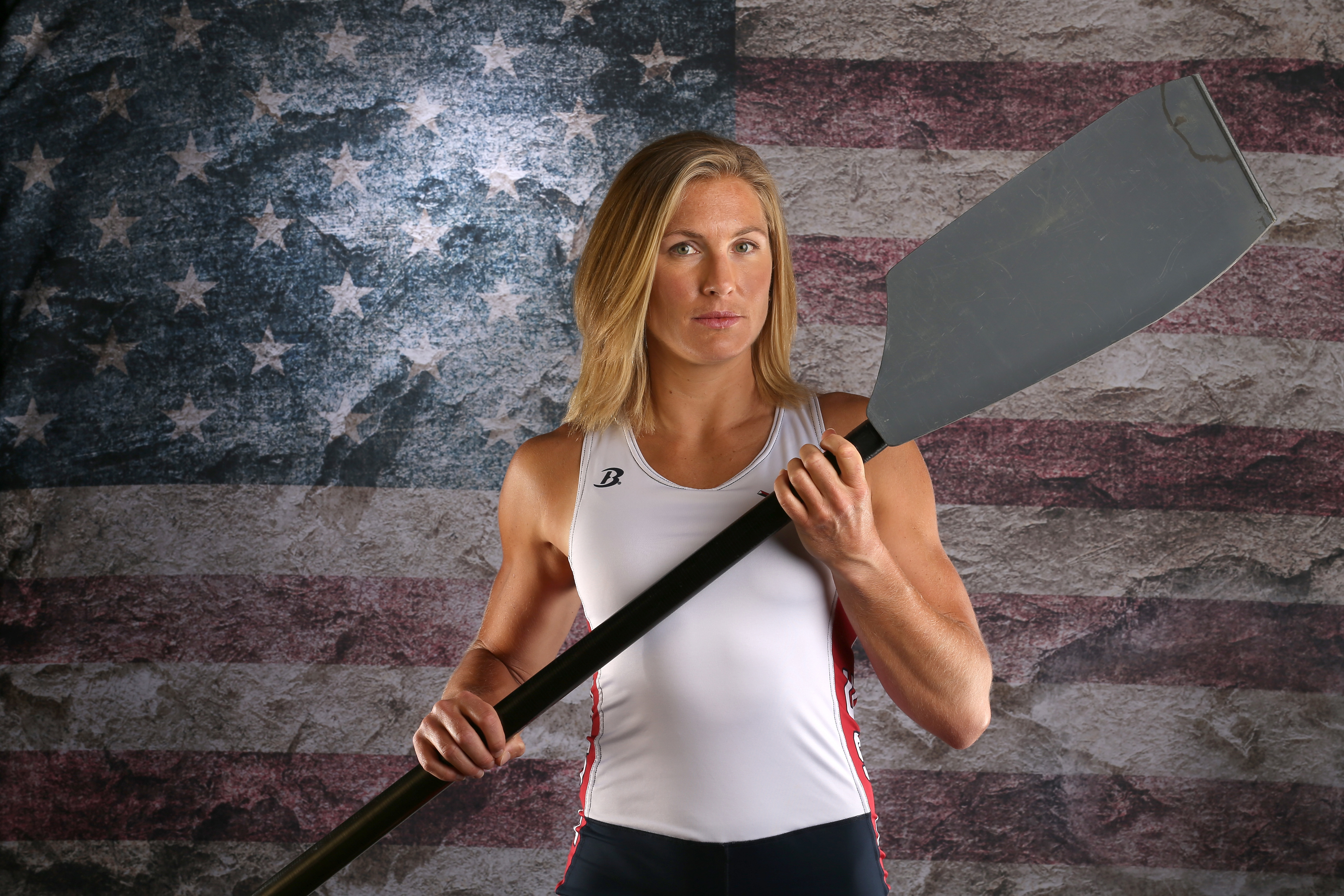Olympic rower Meghan Musnicki holding an oar stands in front of an American flag backdrop, dressed in athletic attire