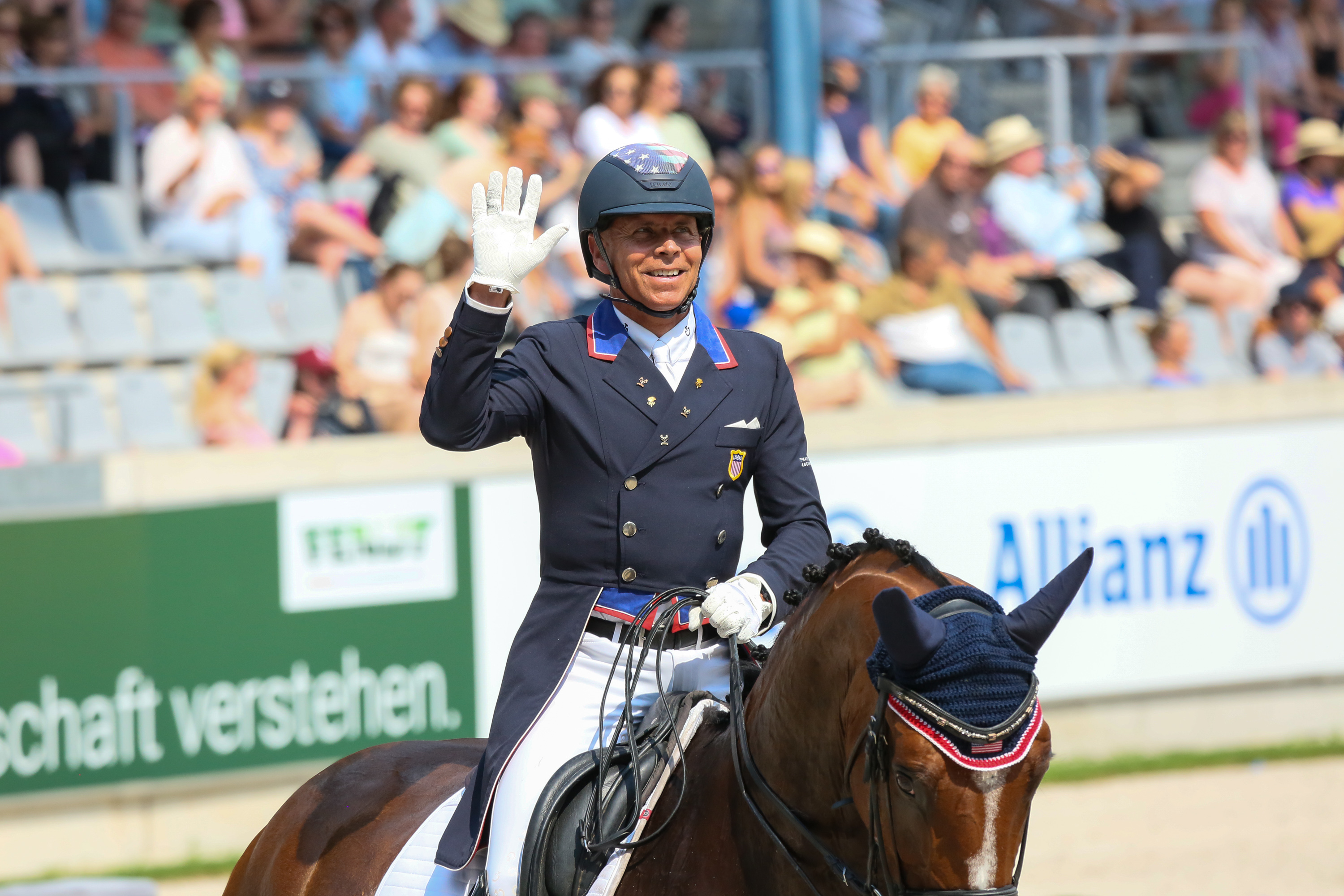 Steffen Peters in formal equestrian attire waves while riding in front of an audience at a sports event