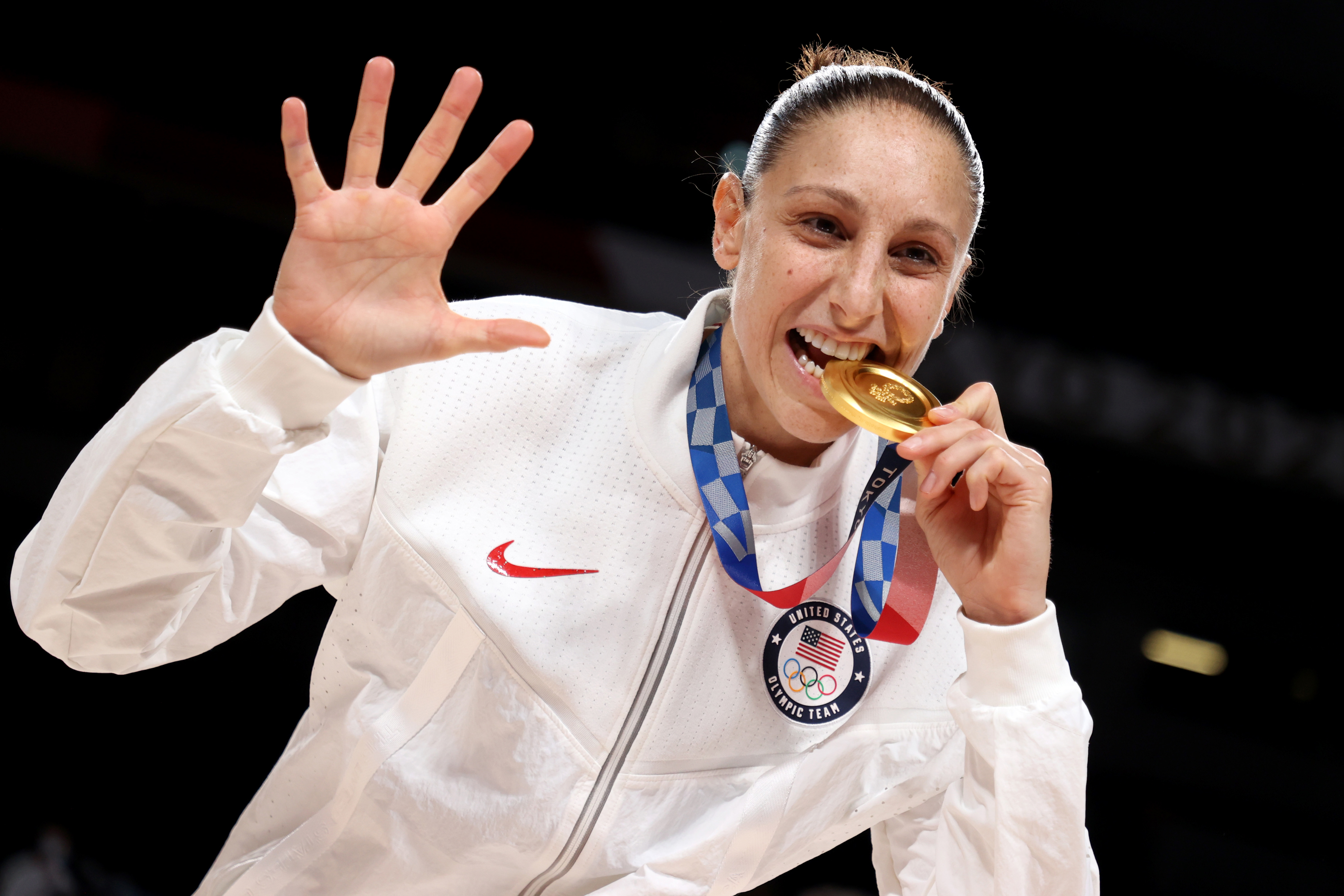 Diana Taurasi, in a white jacket, holds a gold medal with her teeth and raises five fingers, likely indicating her fifth Olympic win, during a sports event