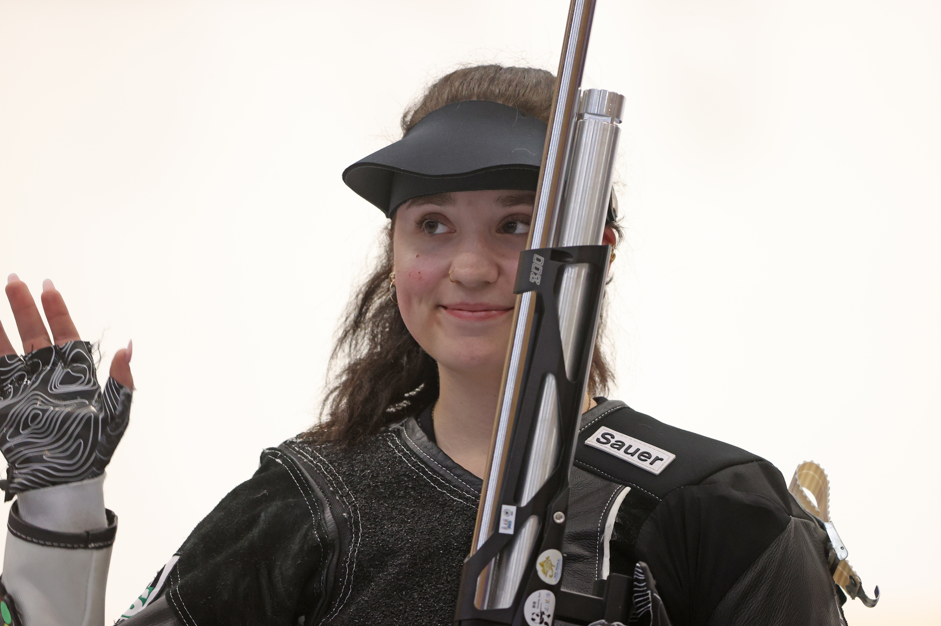 Athlete Mary Tucker waves while holding a rifle, smiling and wearing shooting gear with a visor