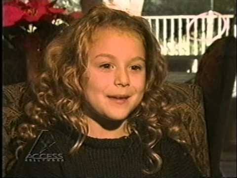 A young child with curly hair, wearing a dark top, is sitting and speaking in an interview on Access Hollywood