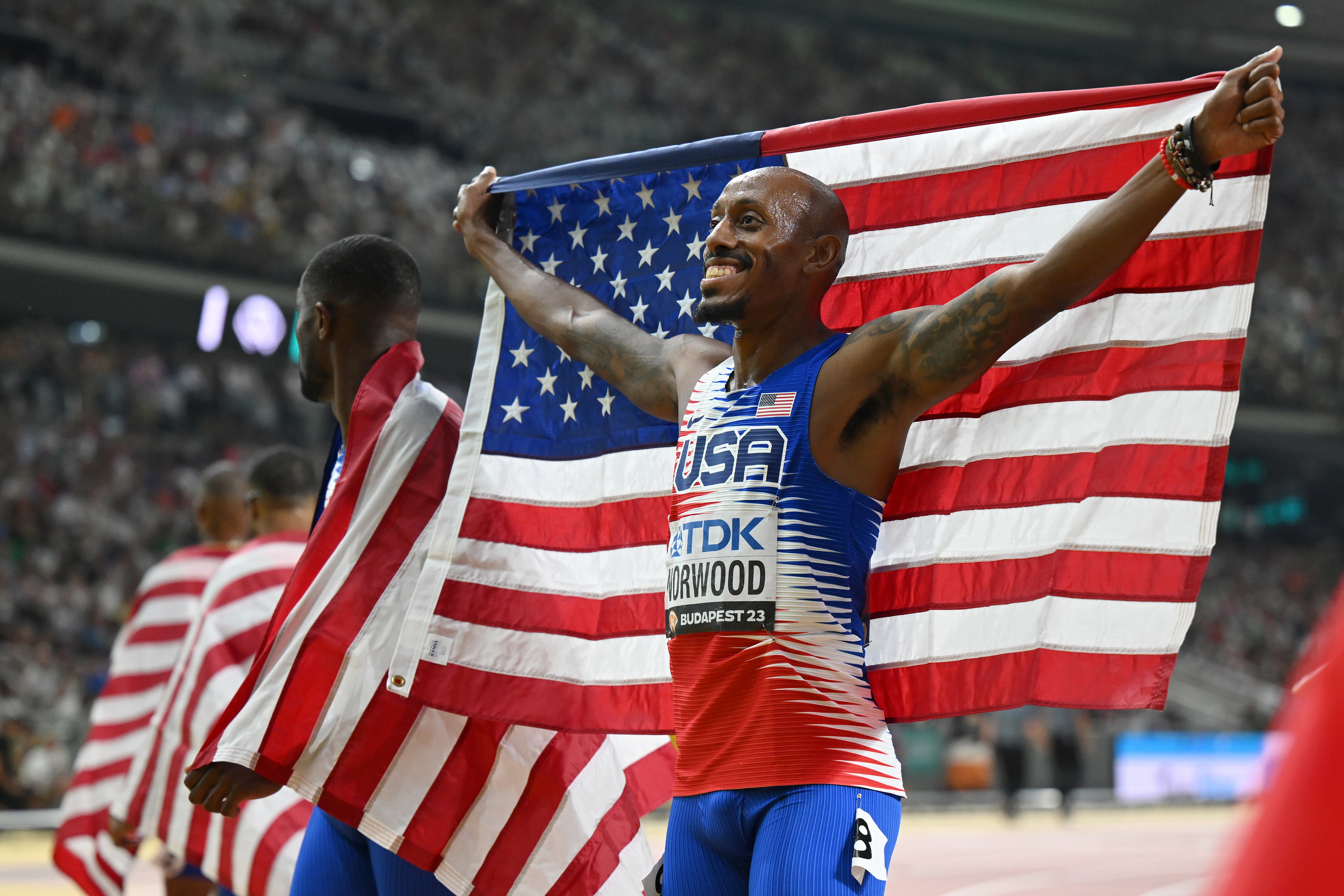 Athletes celebrate with American flags at a sports event, standing in front of a large crowd