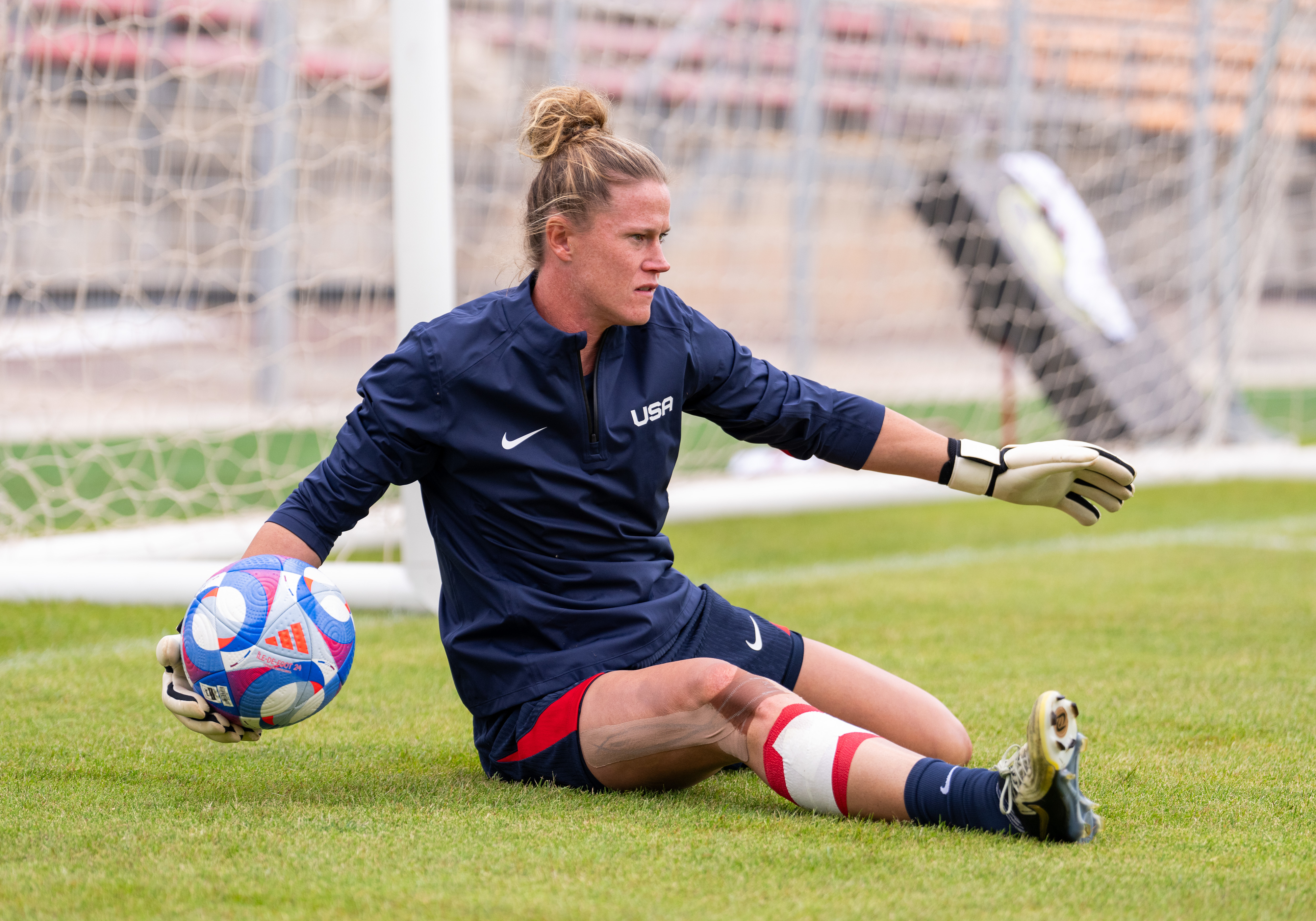 Goalkeeper for the USA women's soccer team, sitting on grass near goalposts, holding a ball, wearing gloves, and sports attire, with a focused expression