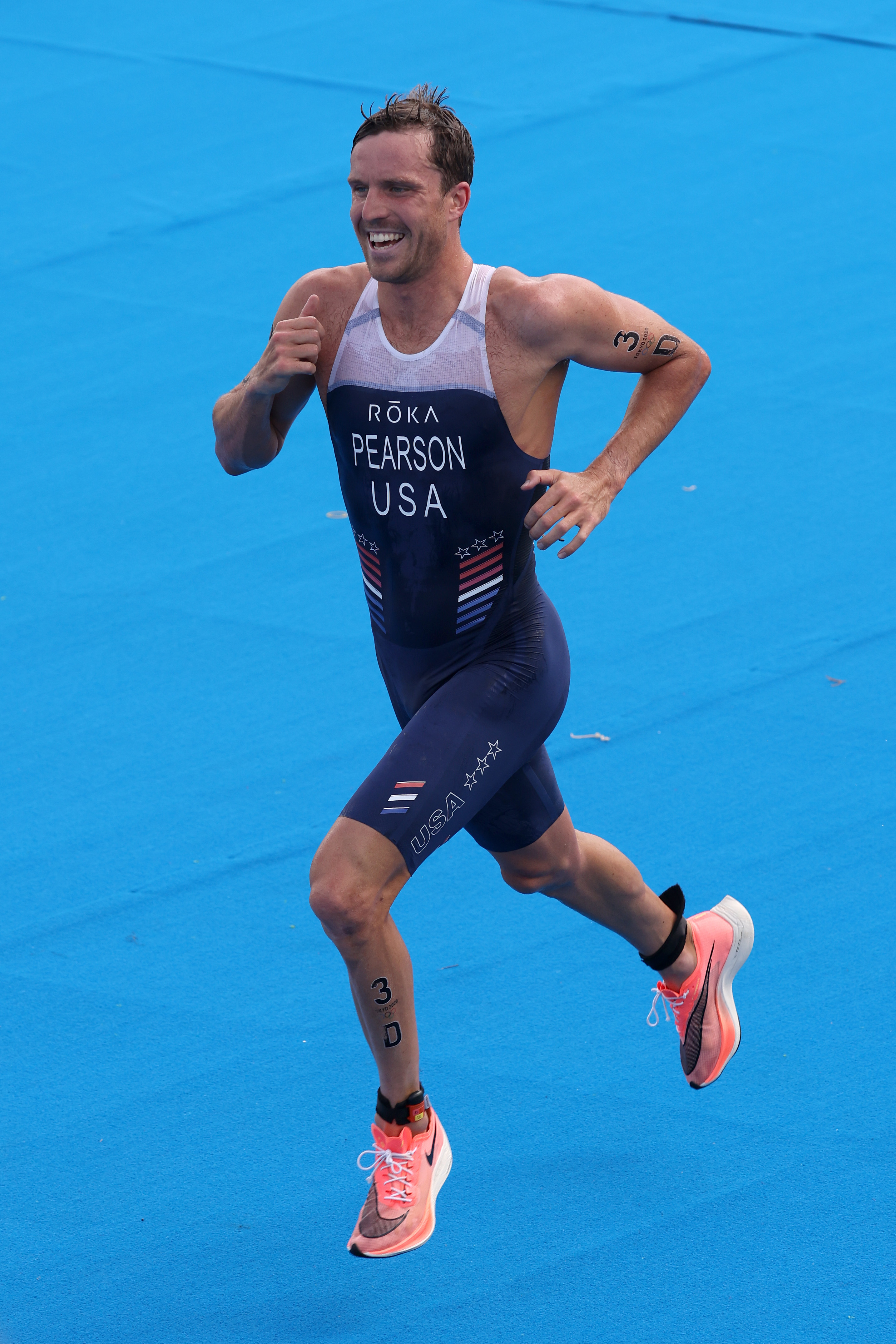 Morgan Pearson running during a triathlon, wearing a dark athletic suit with &quot;Pearson USA&quot; and &quot;Roka&quot; printed on it, smiling and giving a thumbs-up