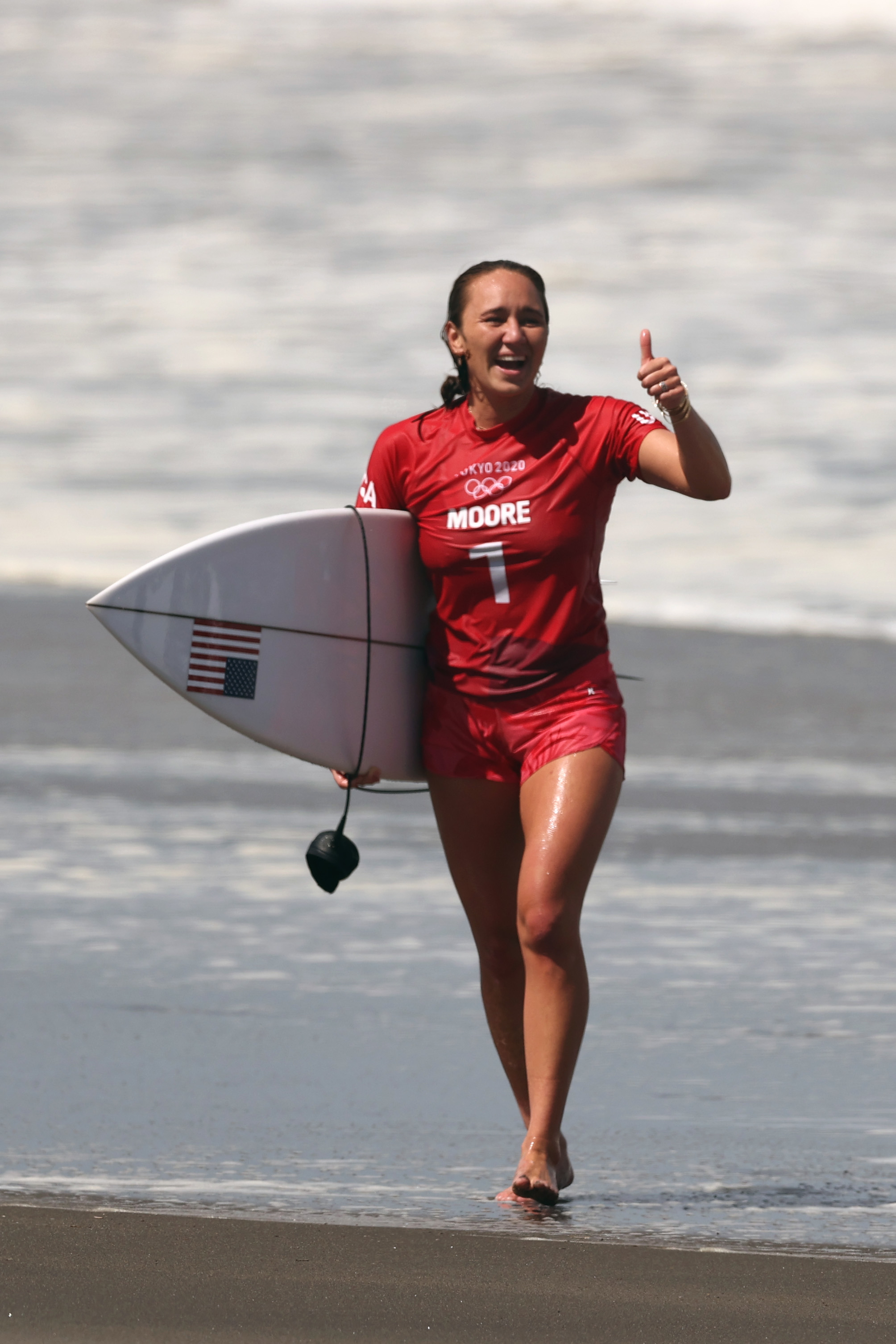 Carissa Moore on the beach holding a surfboard, making a thumbs-up gesture, wearing a sporty outfit