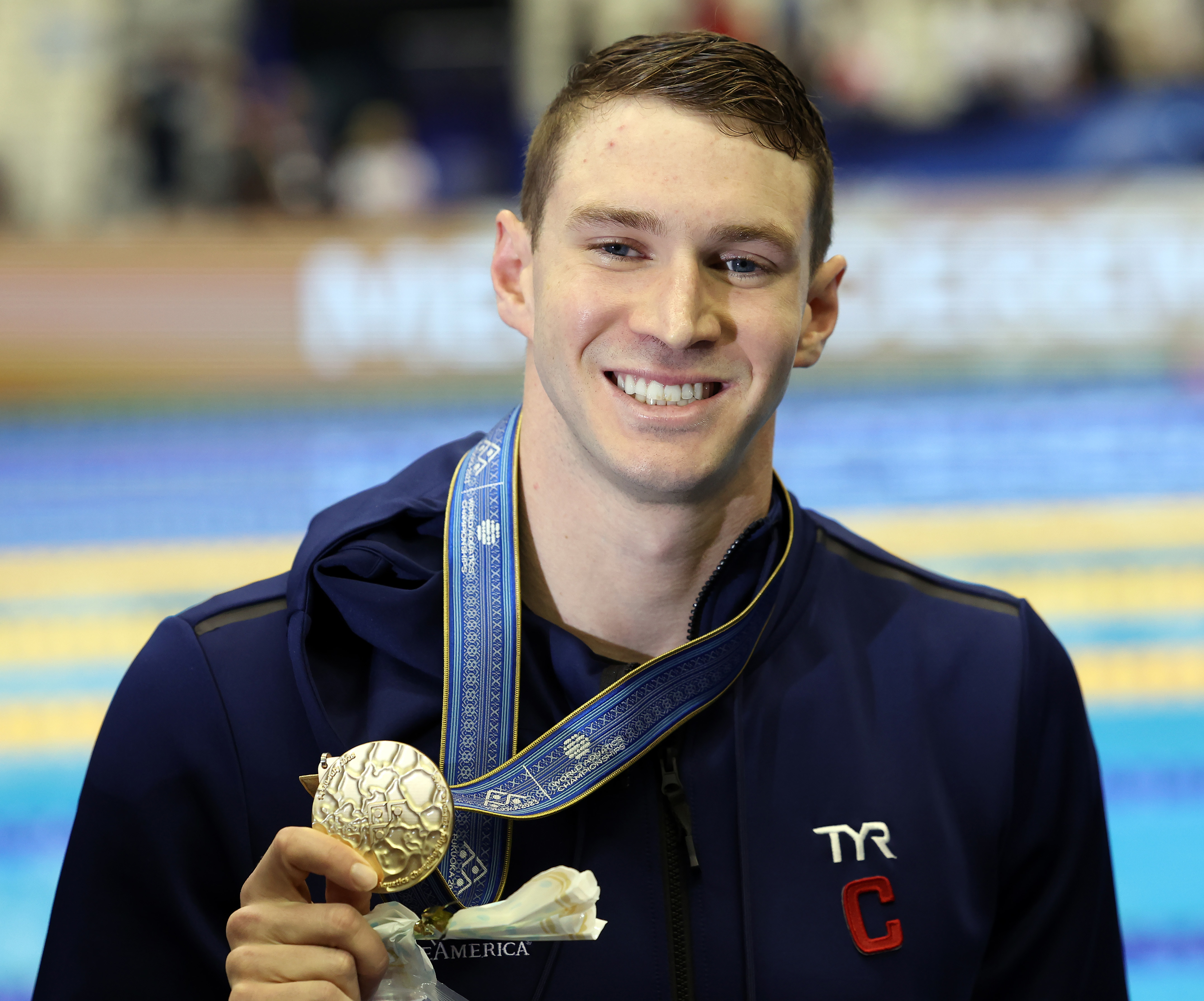 A swimmer stands smiling, holding a gold medal around his neck, dressed in a navy tracksuit