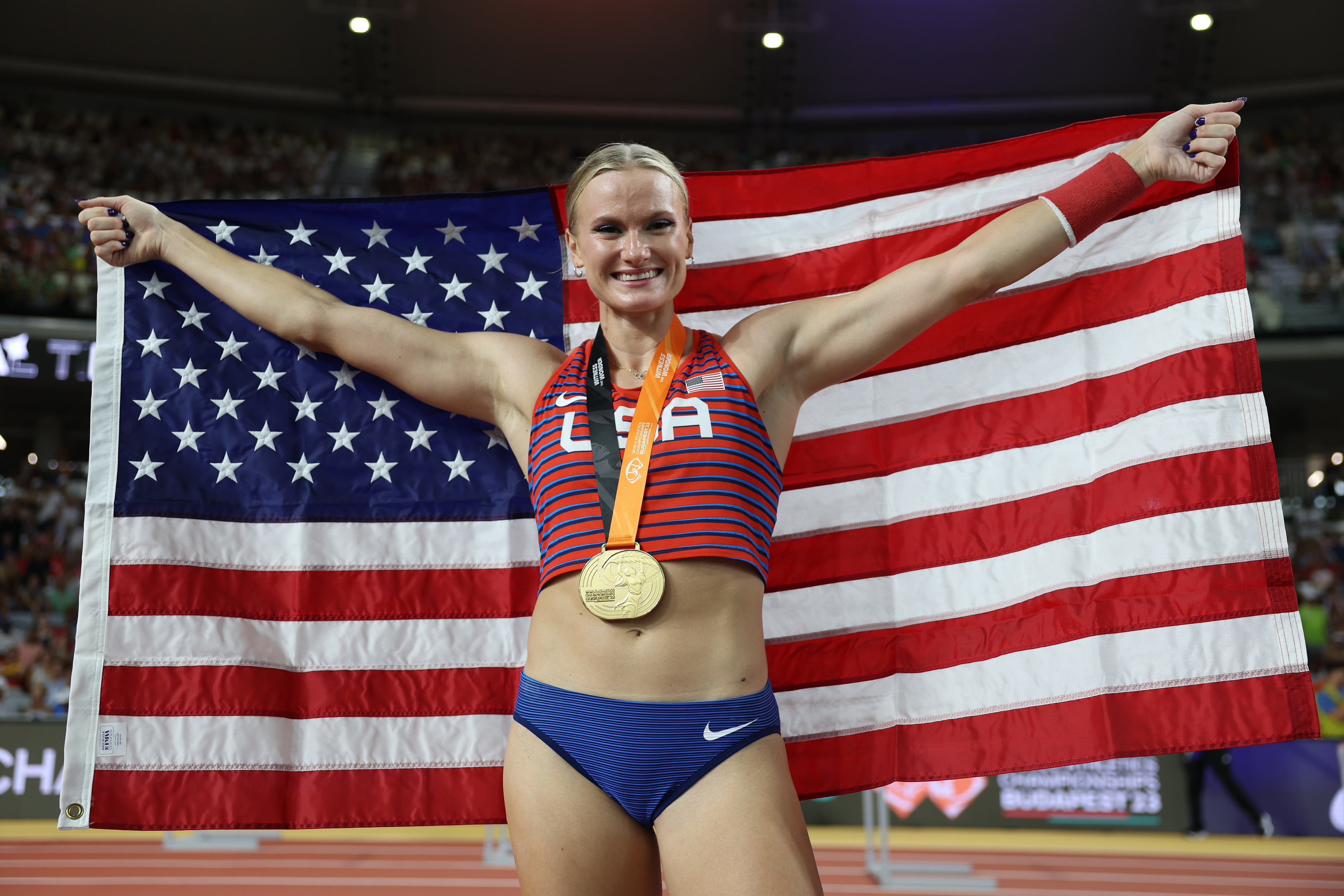 Katie Moon holds the American flag and wears a gold medal around their neck, smiling proudly on a sports field