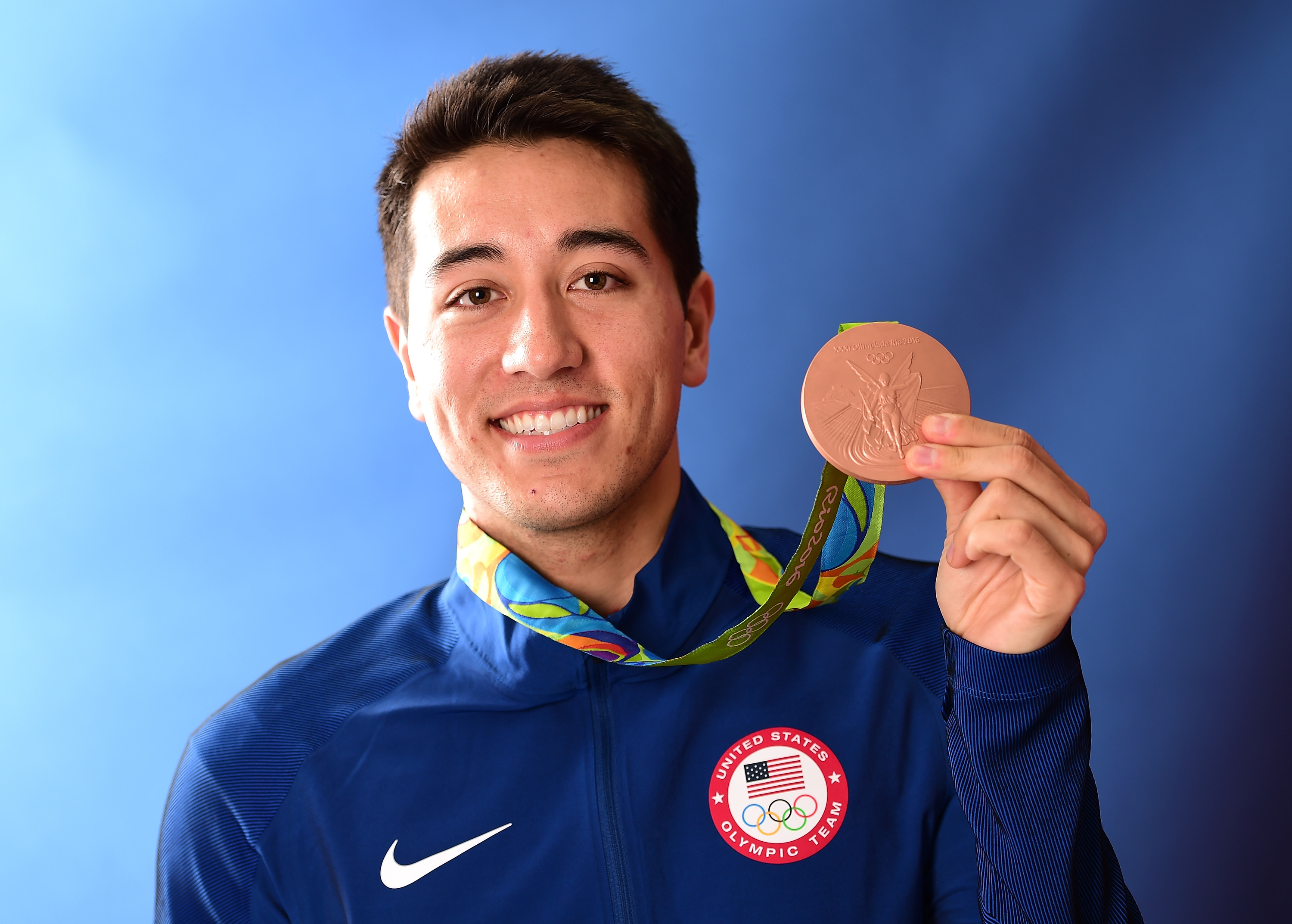 Gerek Meinhardt smiling and holding up his Olympic bronze medal while wearing a Team USA jacket