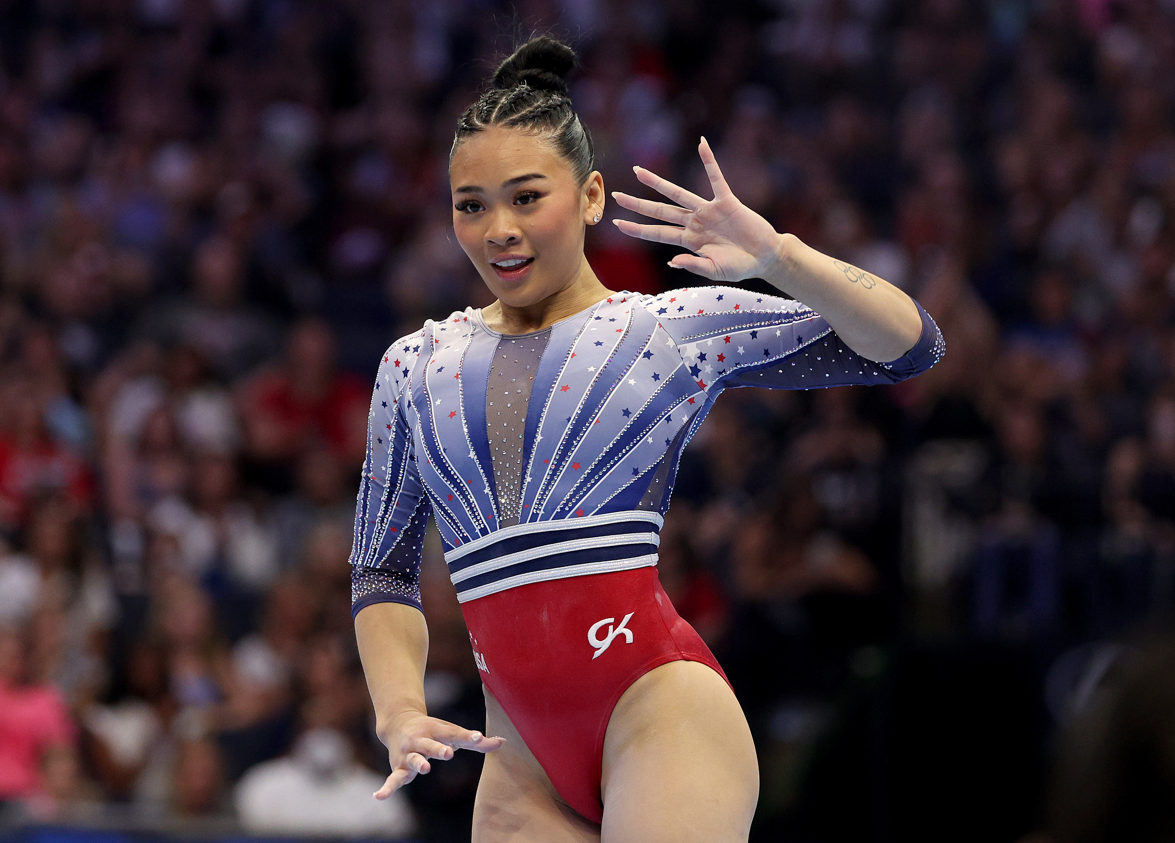 Gymnast Suni Lee performs a floor routine at a competition, wearing a sparkly leotard with a white, blue, and red design