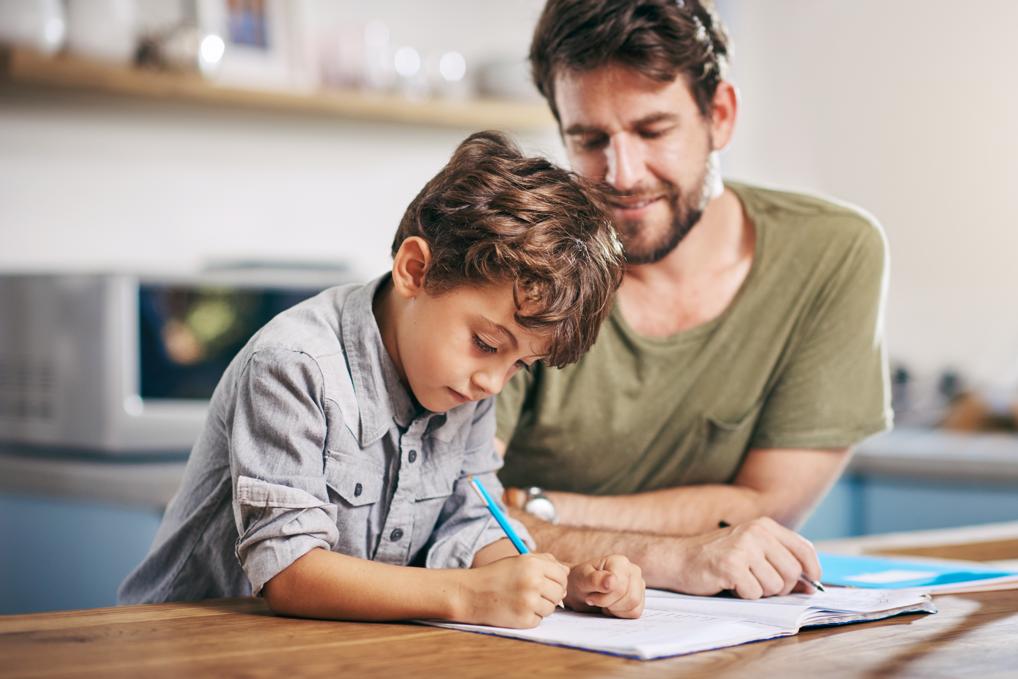 A father with a beard, wearing a casual shirt, helps his young son with curly hair, who is writing in a notebook at a kitchen table