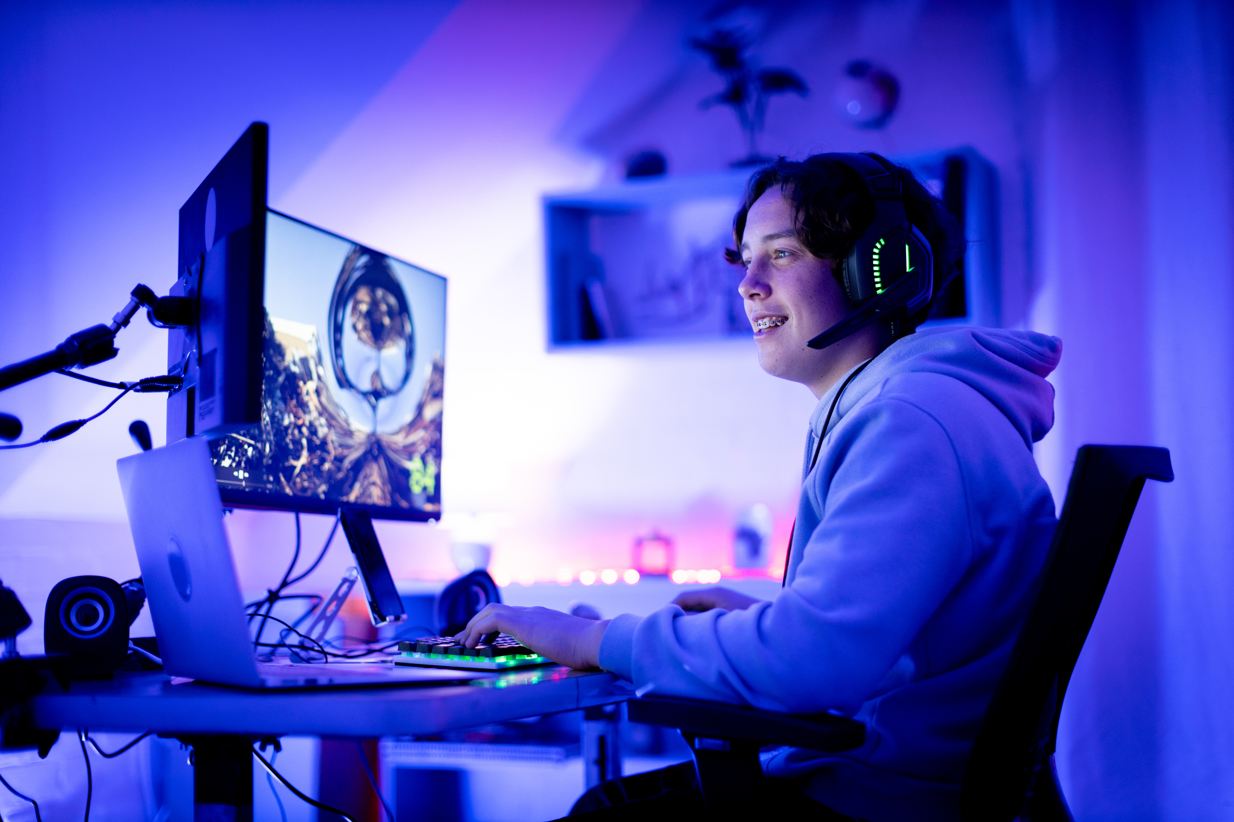 A person wearing a hoodie and headphones is sitting at a desk, engaged in playing a video game on a computer with multiple screens