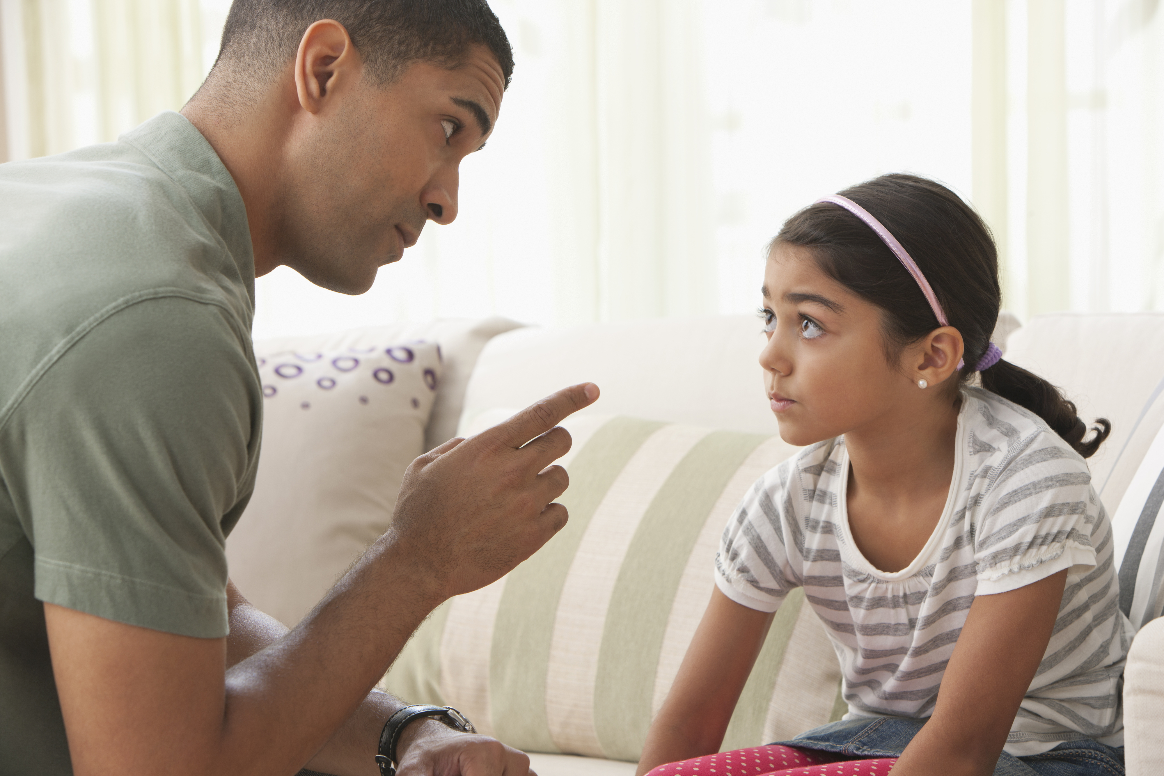 A father gently disciplines his young daughter who sits on a couch, looking attentive