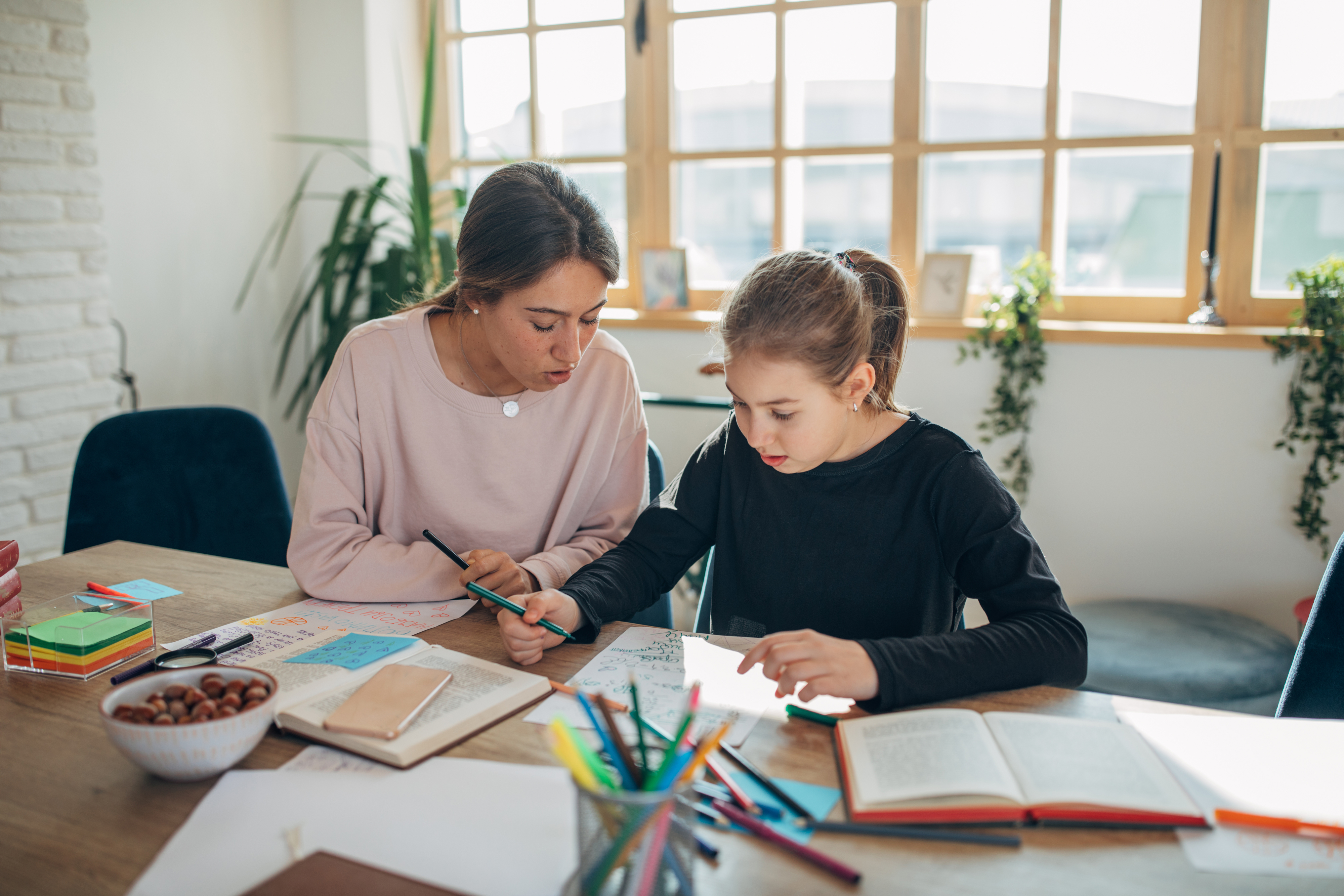 A woman and a young girl study together at a table covered with books, notebooks, and stationery in a well-lit room with large windows