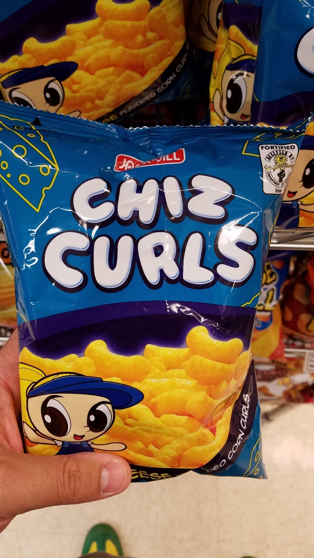 A close-up of a hand holding a bag of Jack ‘n Jill Chiz Curls, a cheese-flavored corn snack featuring a mascot with cartoon eyes and a cap on the packaging