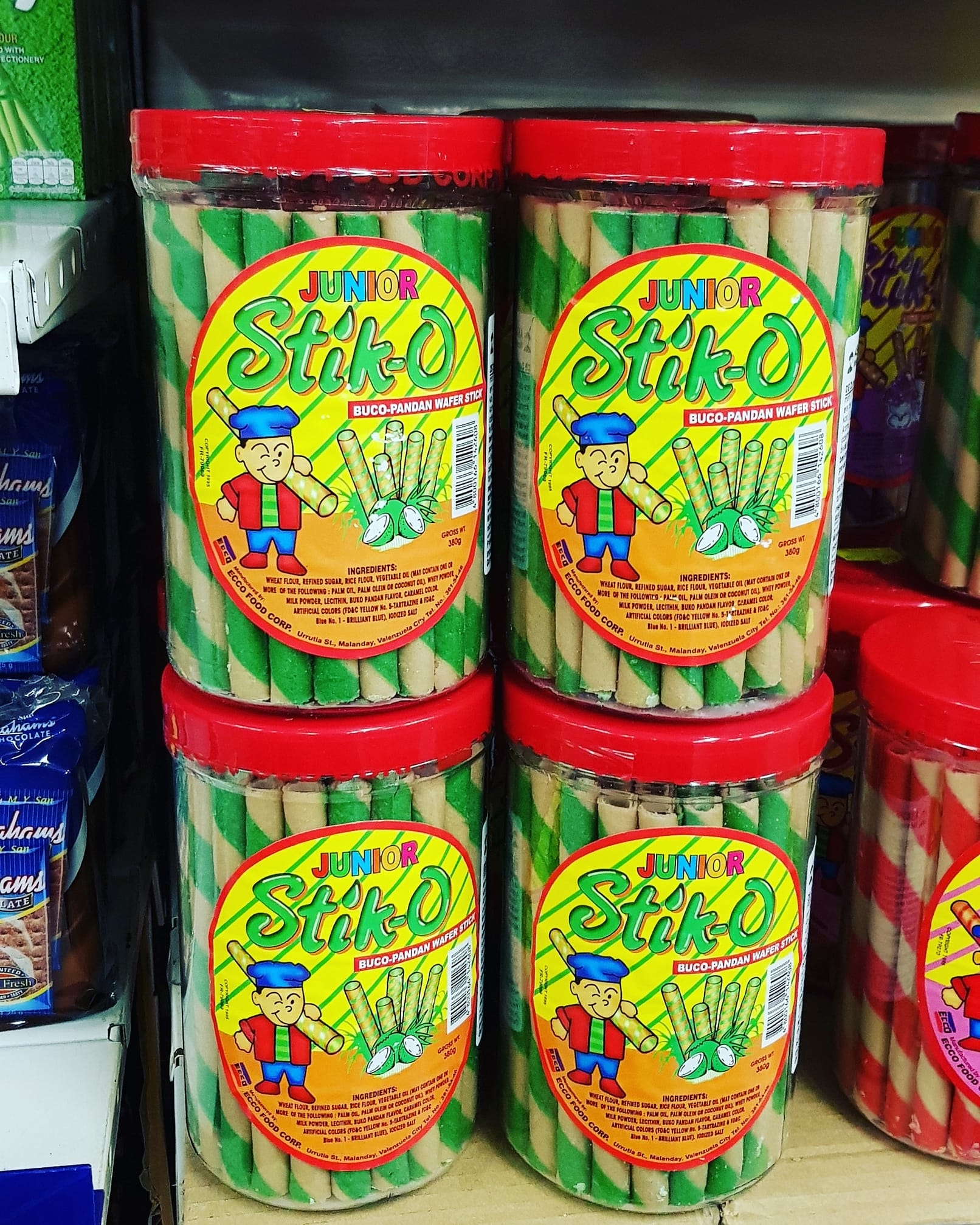 Four containers of Junior Stik-O wafer sticks are displayed on a shelf. The packaging features a cartoon character and images of wafer sticks