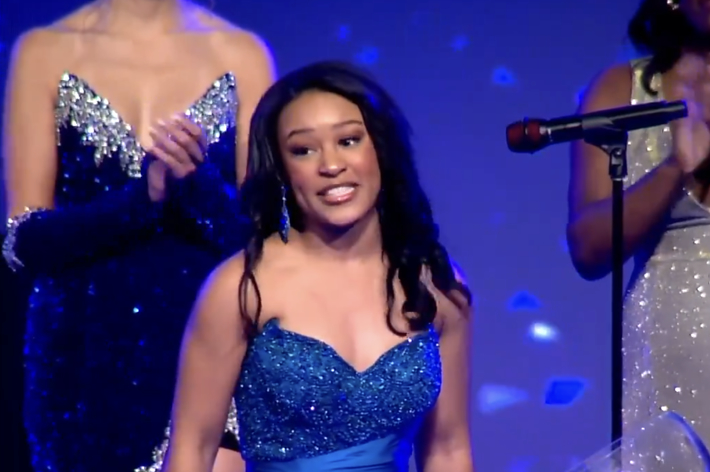 女性が輝く青いドレスを着てスピーチをしている。後方には豪華なドレスを着た女性が拍手を送っている。