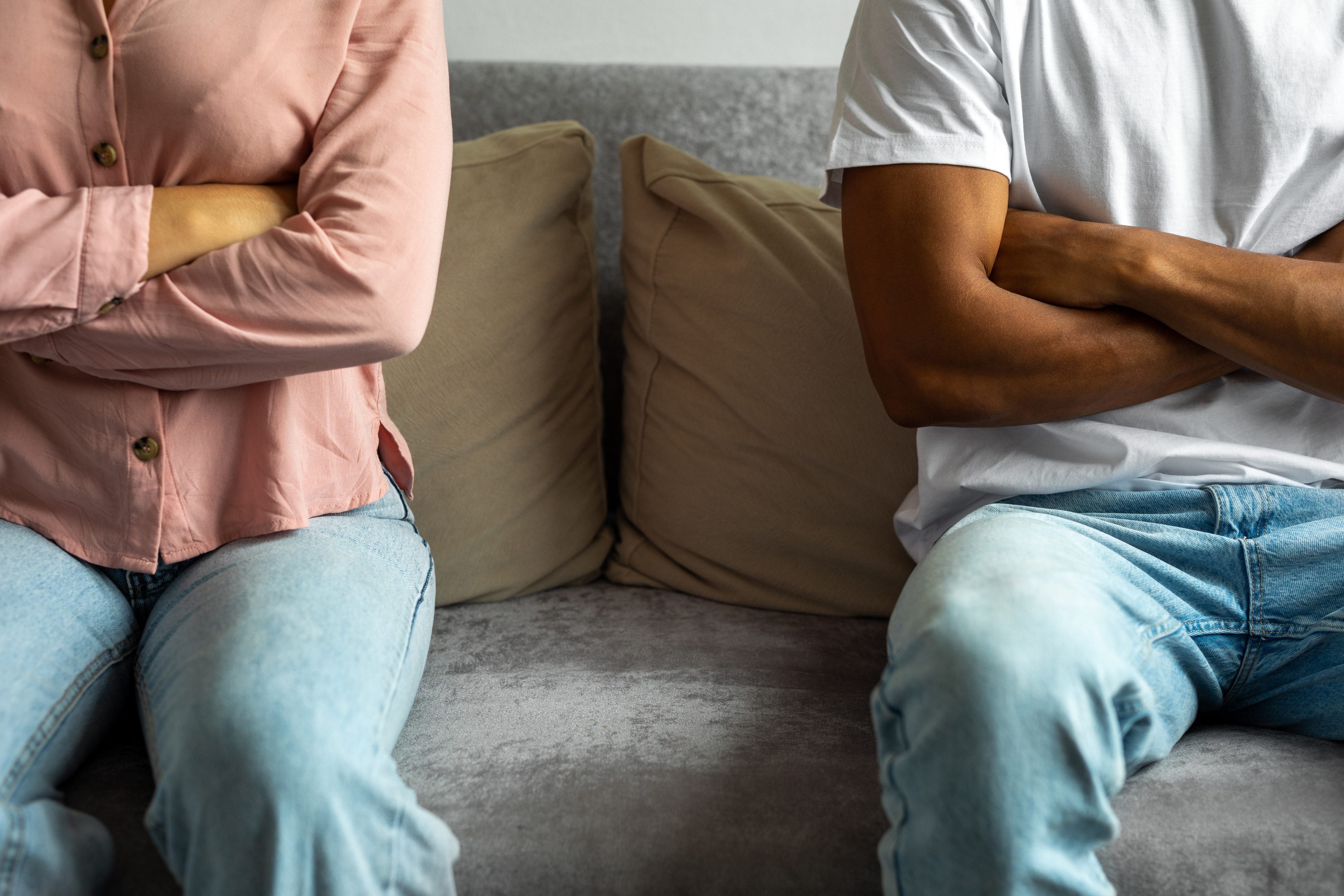 Two people sitting on a sofa with their arms crossed, facing away from each other, showing signs of disagreement. Their faces are not visible