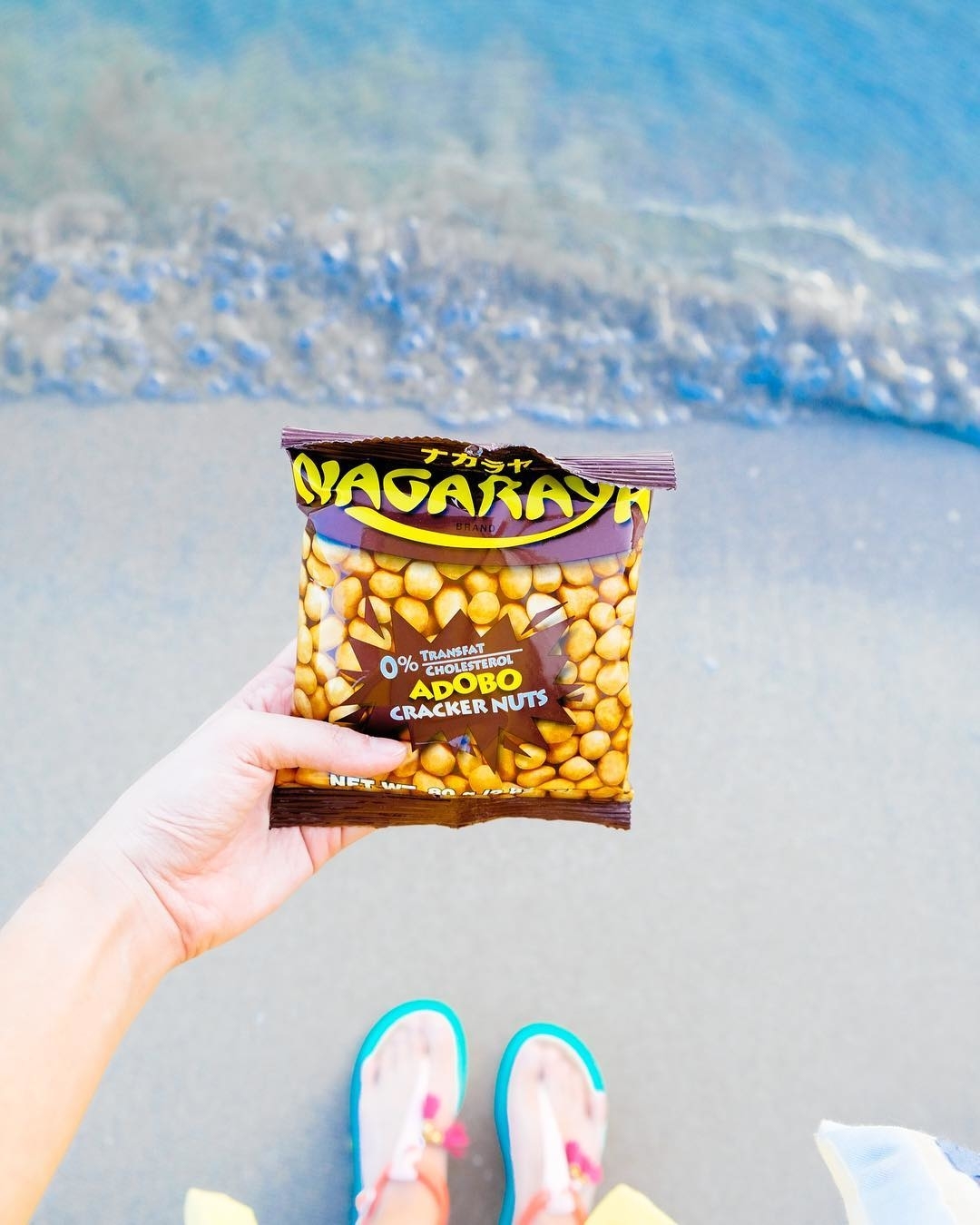 Bag of Oishi Nagaraya Adobo Cracker Nuts held over sandy beach with water in the background