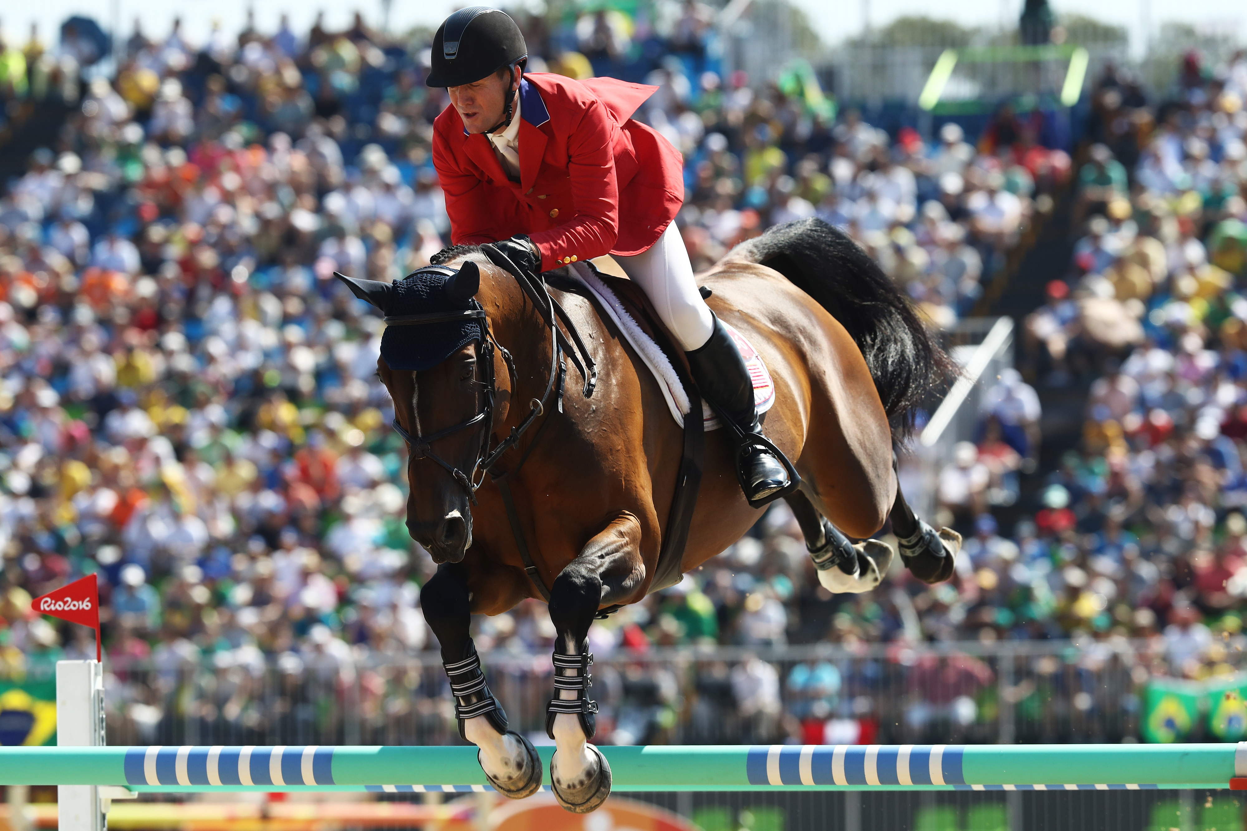 An equestrian rider wearing a red blazer and black helmet jumps a horse over an obstacle during a competitive event