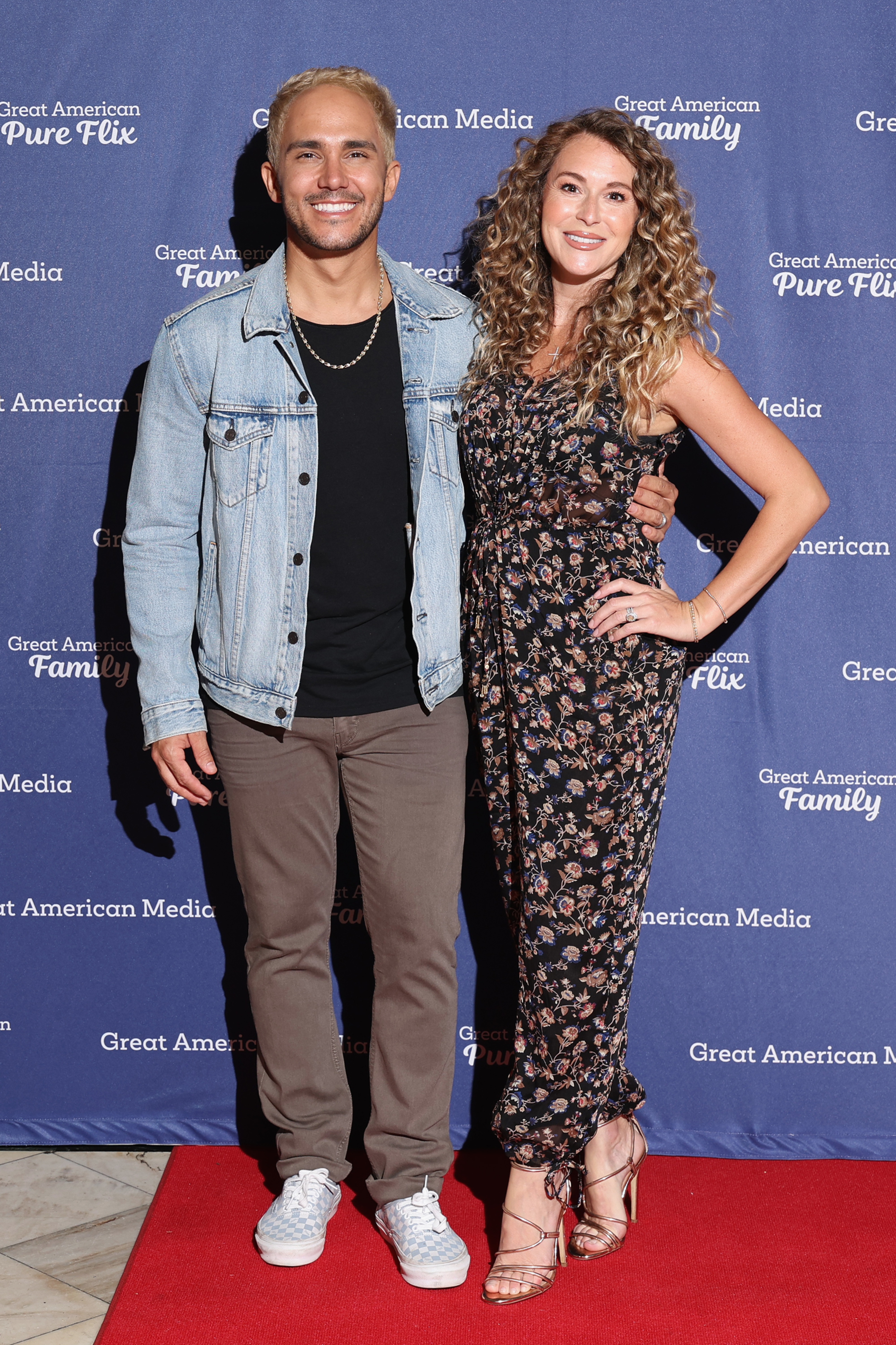 Carlos PenaVega in a denim jacket and Alexa PenaVega in a floral dress pose together on a red carpet at a Great American Family event