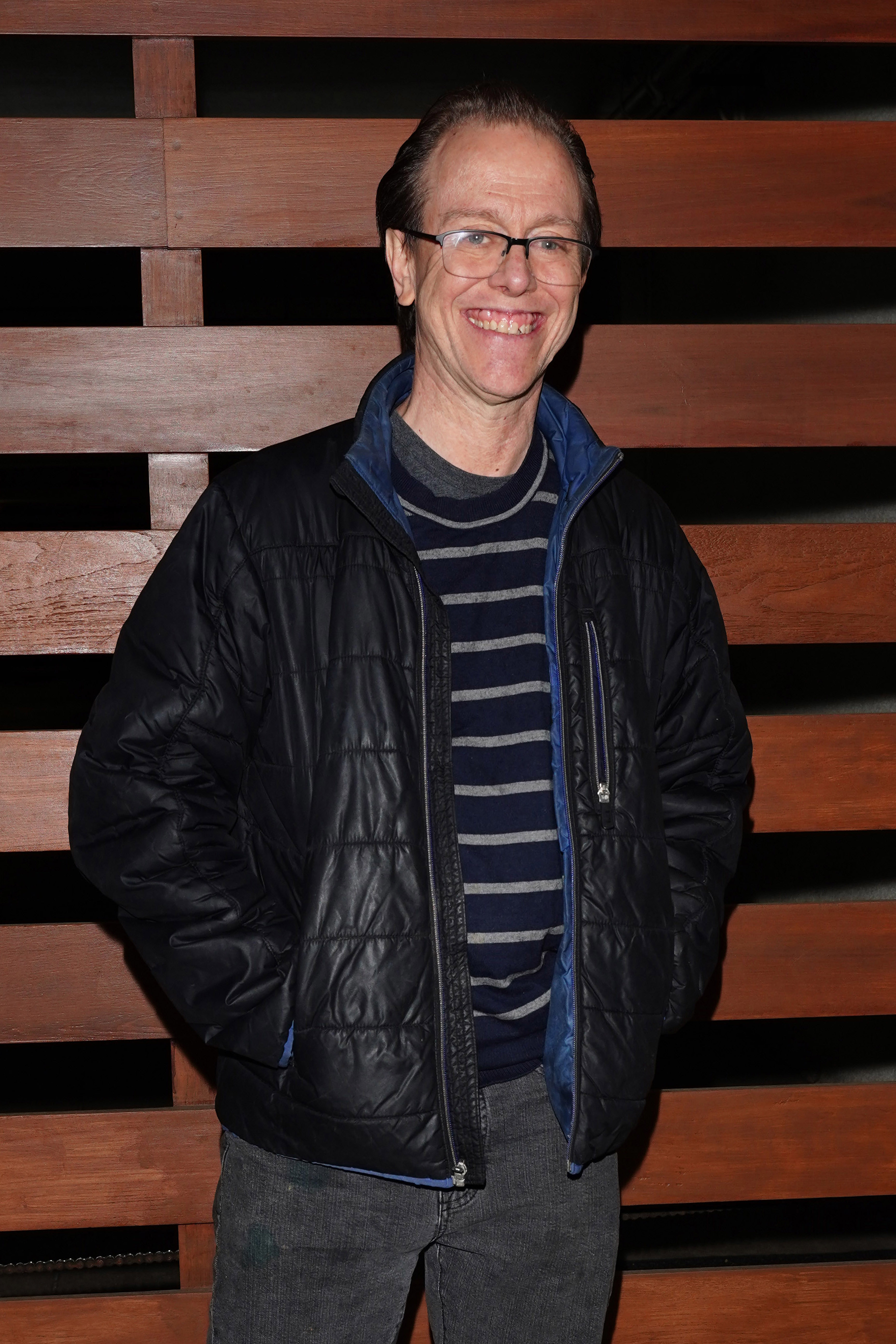 Sean Whalen smiling with hands in pockets wearing glasses, a striped sweater, and a puffy jacket standing in front of a wooden backdrop