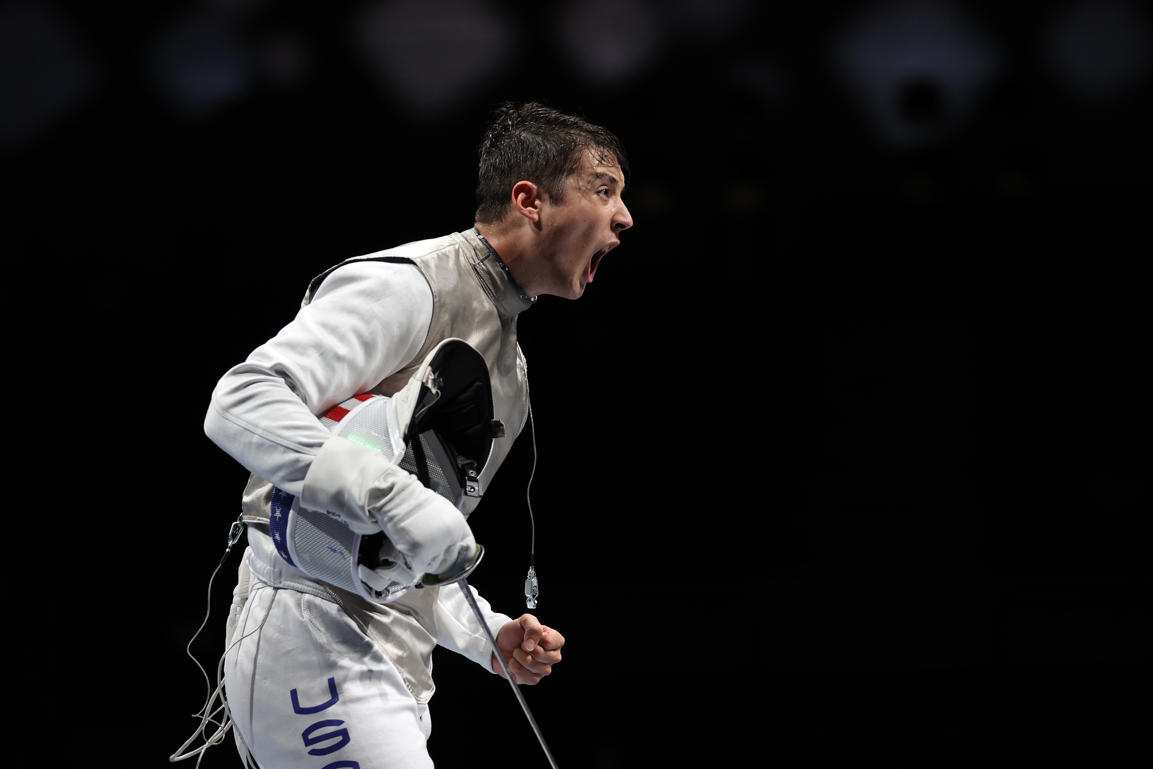 A fencer, clad in full protective gear, passionately celebrates a victory during a match. The letters &quot;USA&quot; are visible on their pants