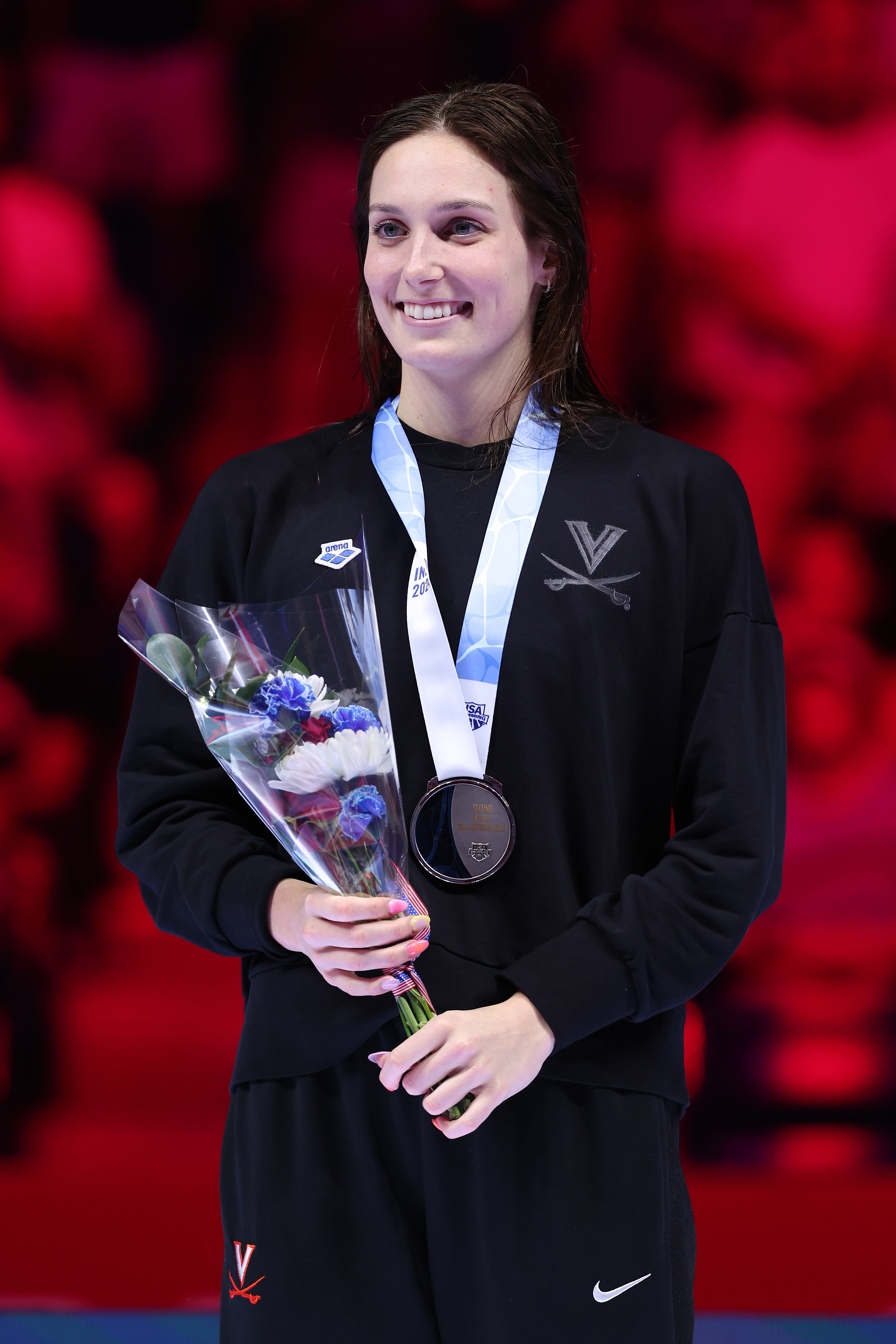 A female athlete in a sports tracksuit holds a bouquet and wears a silver medal around her neck at an award ceremony