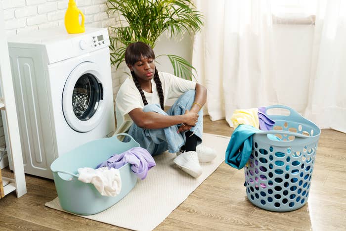 A person sits on the floor next to a washing machine, surrounded by two laundry baskets filled with clothes