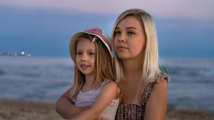 Two unidentified women, one younger and one older, are sitting on a beach. Both have light hair; the younger wears a hat and the older has long, loose hair