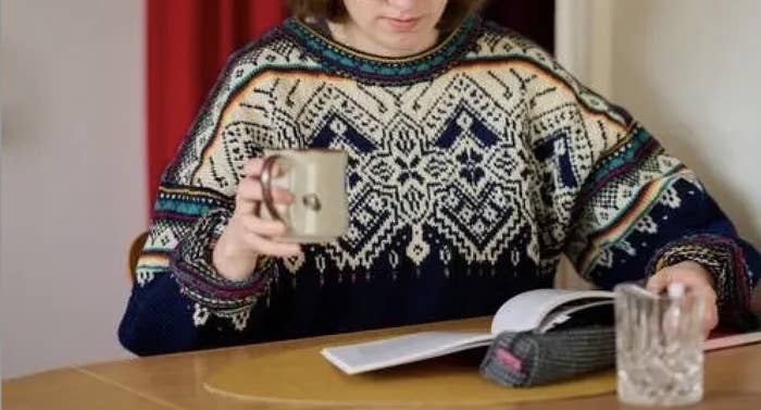 A person in a patterned sweater holds a mug and reads a book on a table
