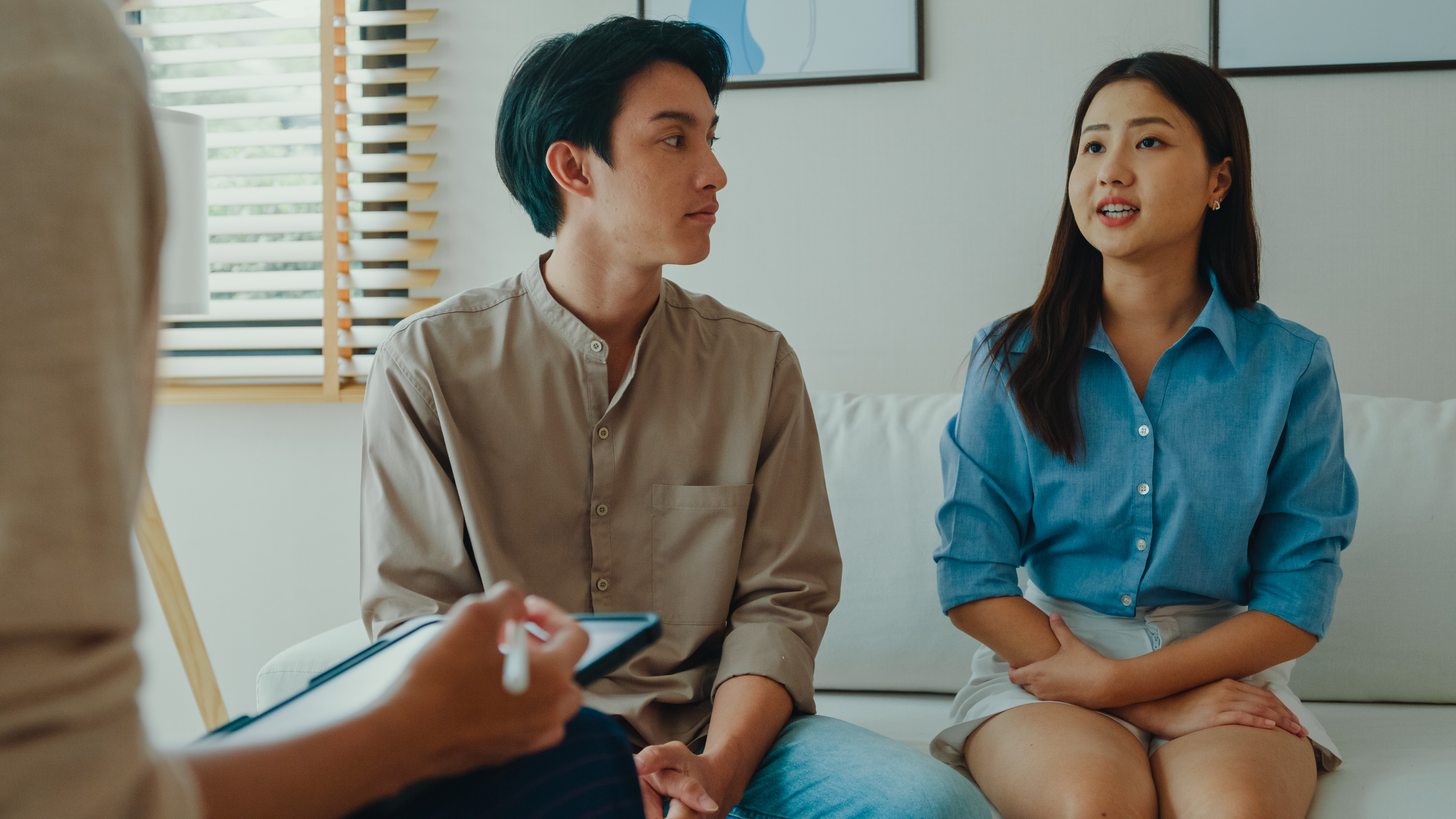 A man and a woman, seated on a couch, engage in conversation with a professional holding a clipboard. The setting suggests a counseling or therapy session