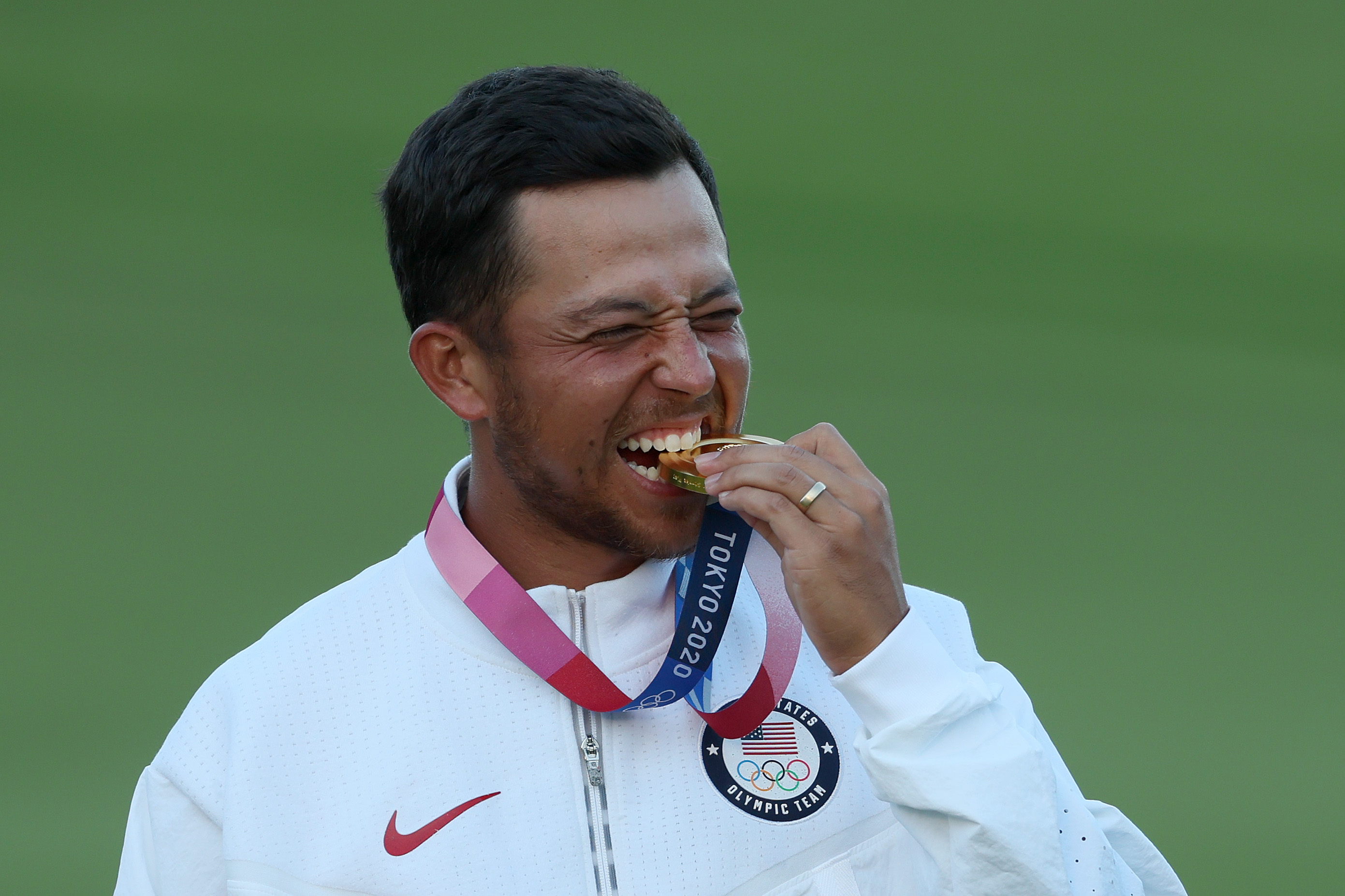 Xander Schauffele, wearing a USA jacket, bites his Tokyo 2020 Olympic gold medal