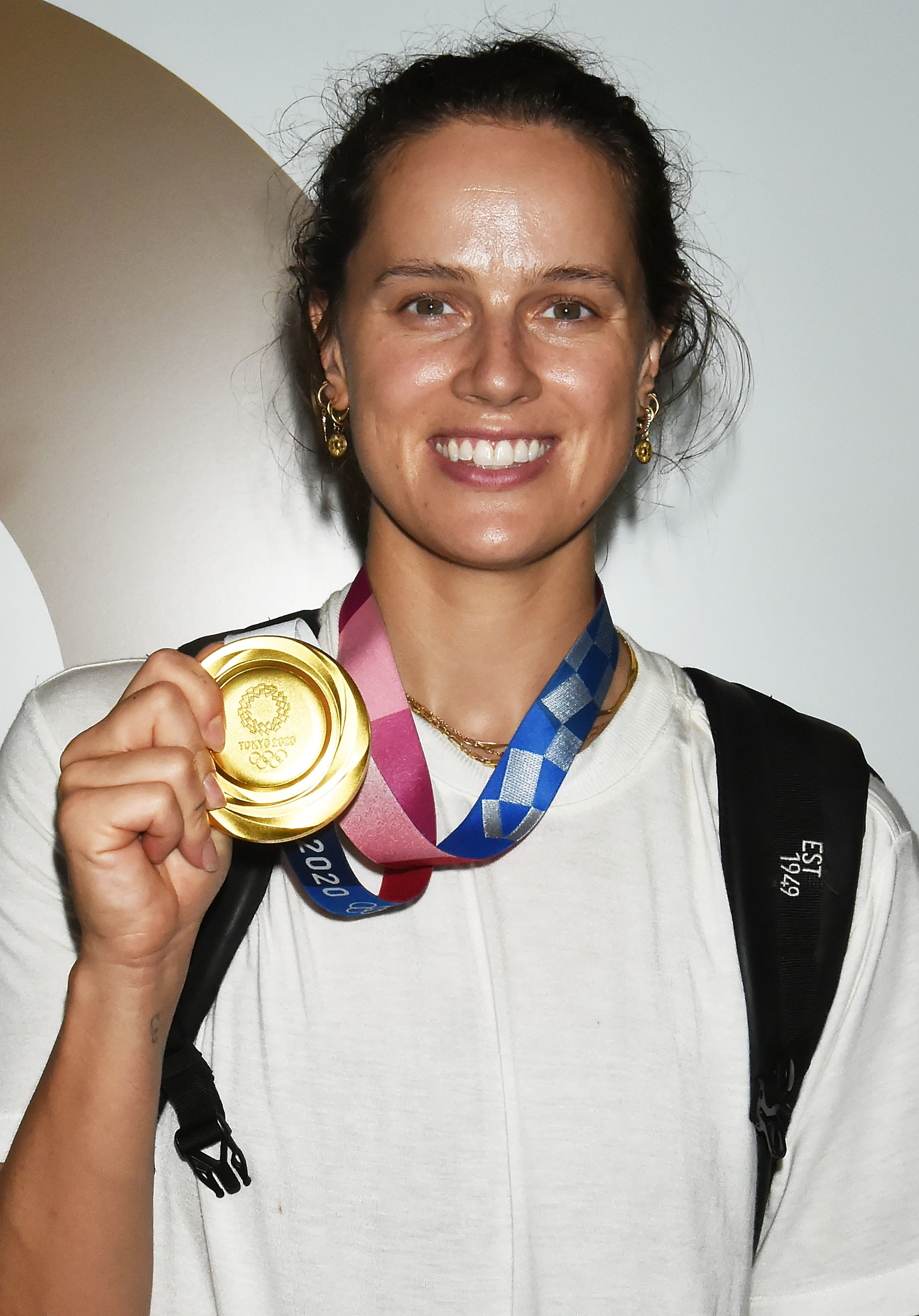 Olympic athlete Ariarne Titmus smiling and holding her gold medal from the 2020 Olympics, wearing a white shirt and a backpack