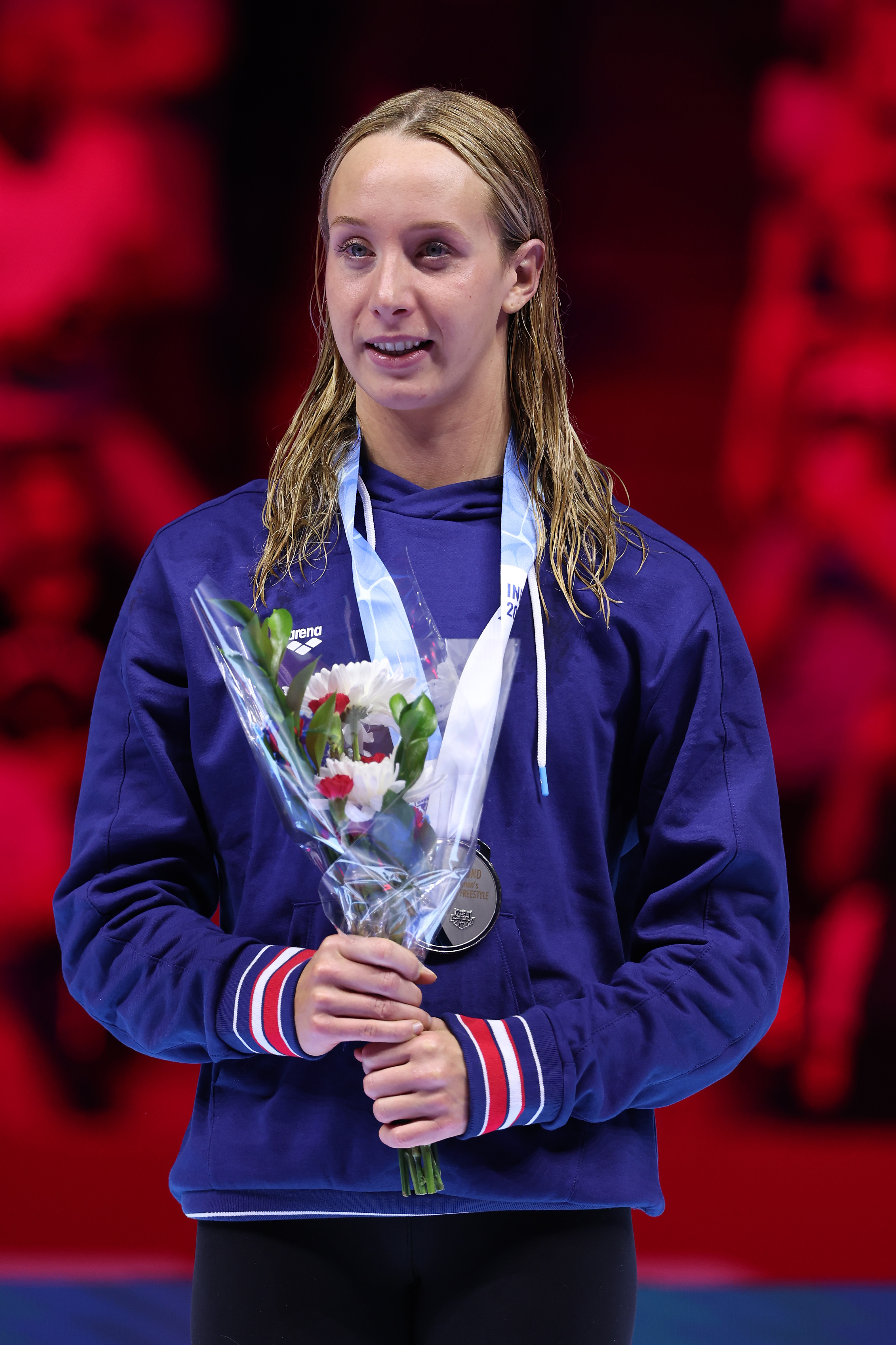 A female athlete stands on an awards stage holding a bouquet and wearing a medal around her neck. She is dressed in a sport jacket