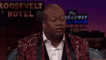 Tituss Burgess, wearing a patterned blazer and white shirt, closes his eyes and takes a deep breath on a talk show set with "Roosevelt Hotel" and "The Broadway Hollywood" signs