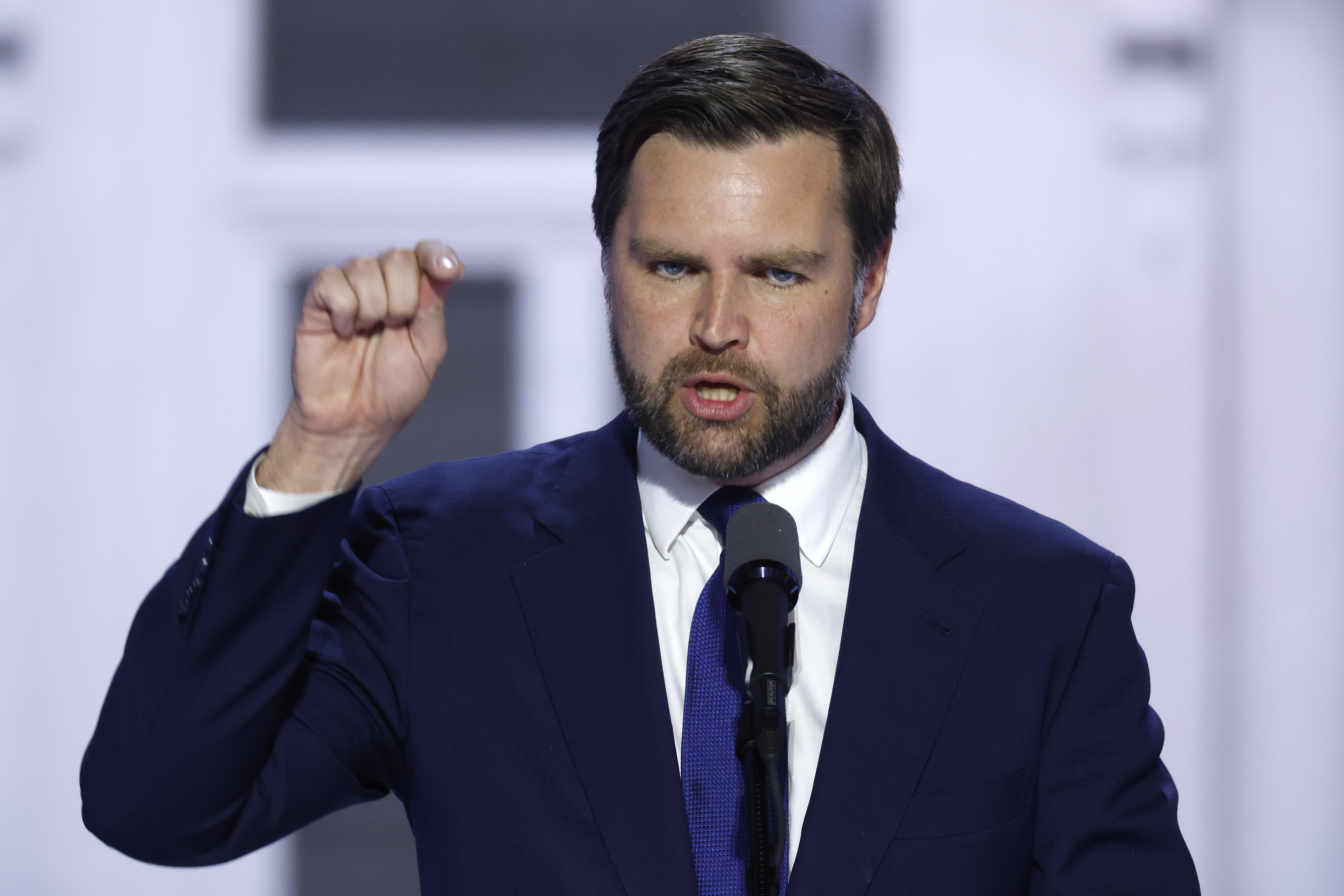 J.D. Vance speaks passionately at a podium, gesturing with his right hand in a blue suit and tie