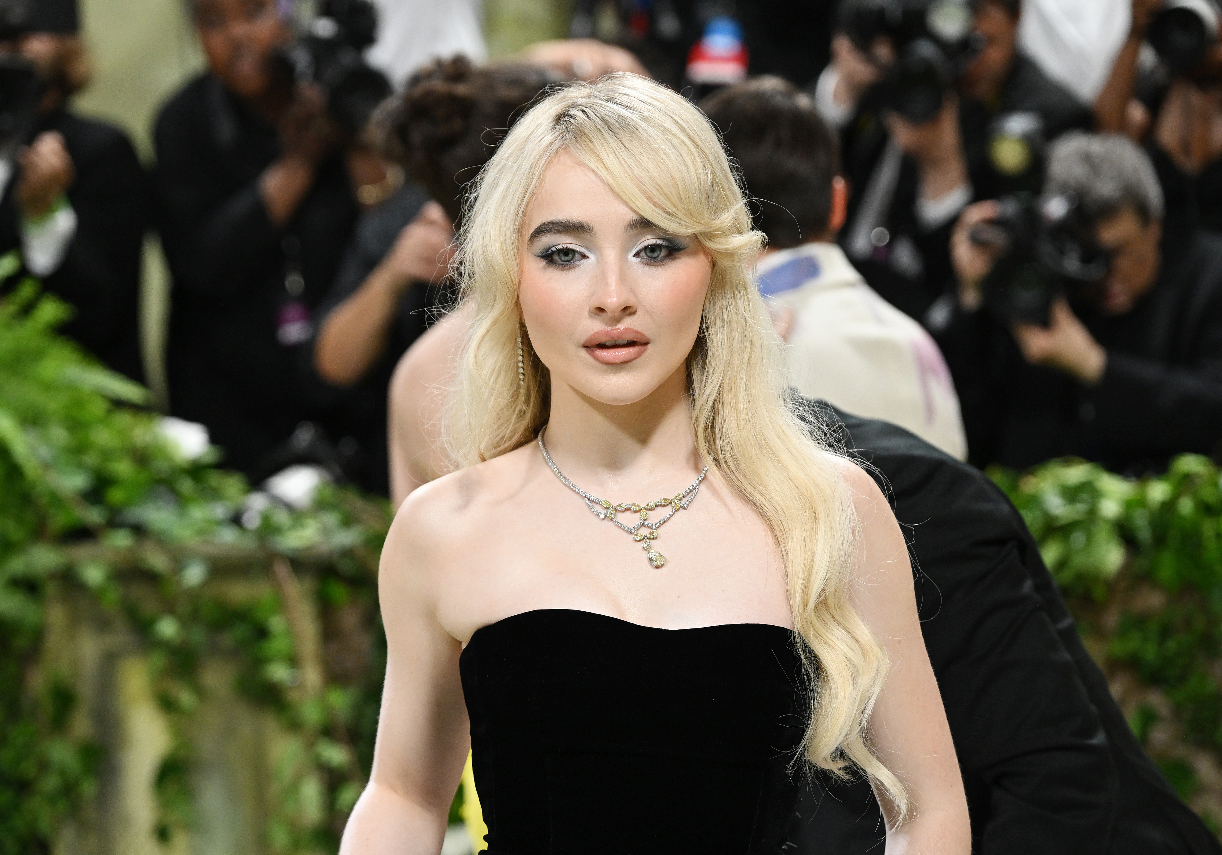 Sabrina Carpenter on a red carpet in an elegant strapless black gown, wearing layered necklaces. A crowd and photographers are in the background