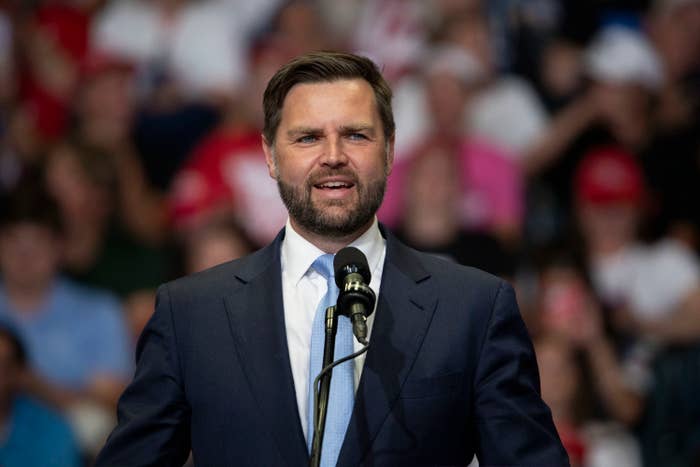 Vance is speaking at a podium in front of a crowd. He is wearing a suit and tie. The background is blurred and shows people in the audience