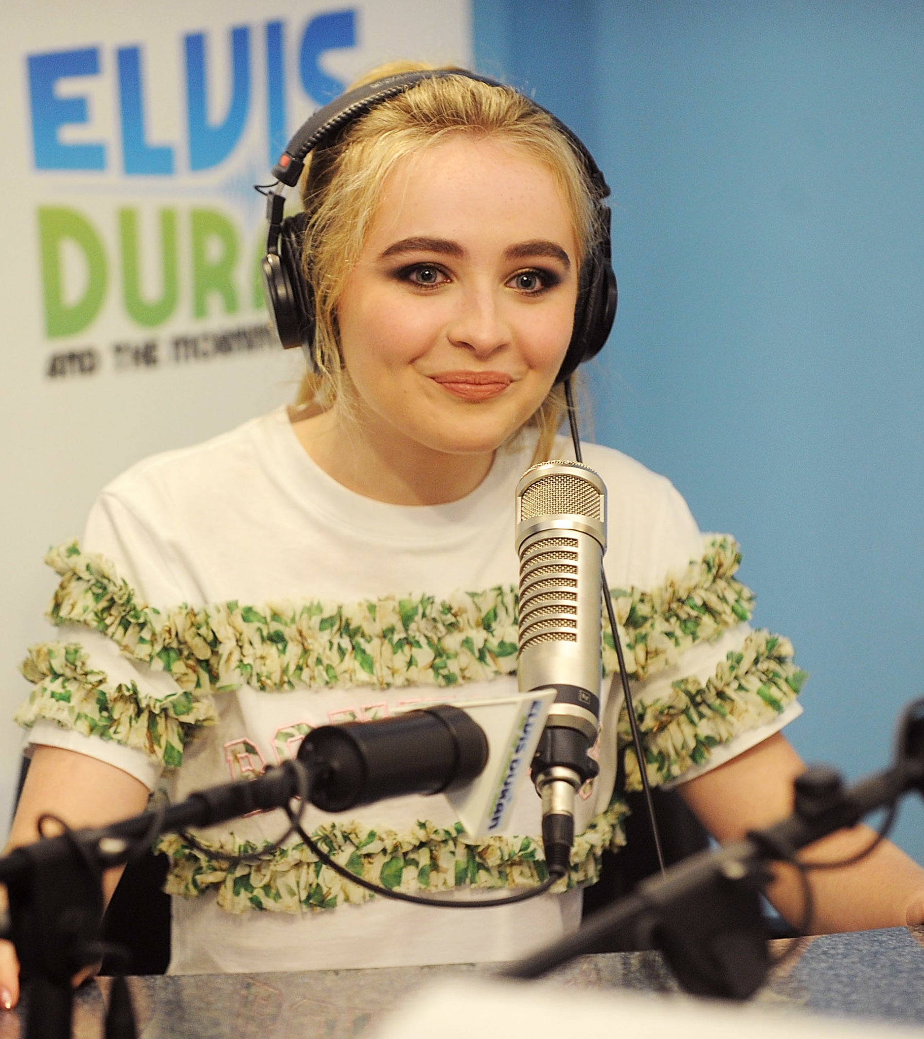 Sabrina Carpenter, wearing a casual top, speaks into a microphone during her appearance on the Elvis Duran and the Morning Show