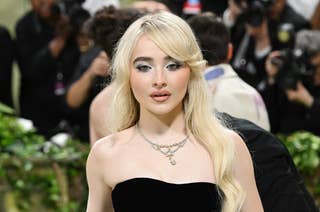 Sabrina Carpenter at a celebrity event, wearing a strapless, elegant black gown and a delicate necklace, with camera crews in the background