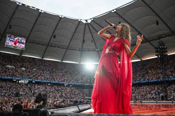 Taylor Swift performing on stage in a stadium, passionately singing into the microphone while wearing an elegantly styled dress. Crowd and screen in background