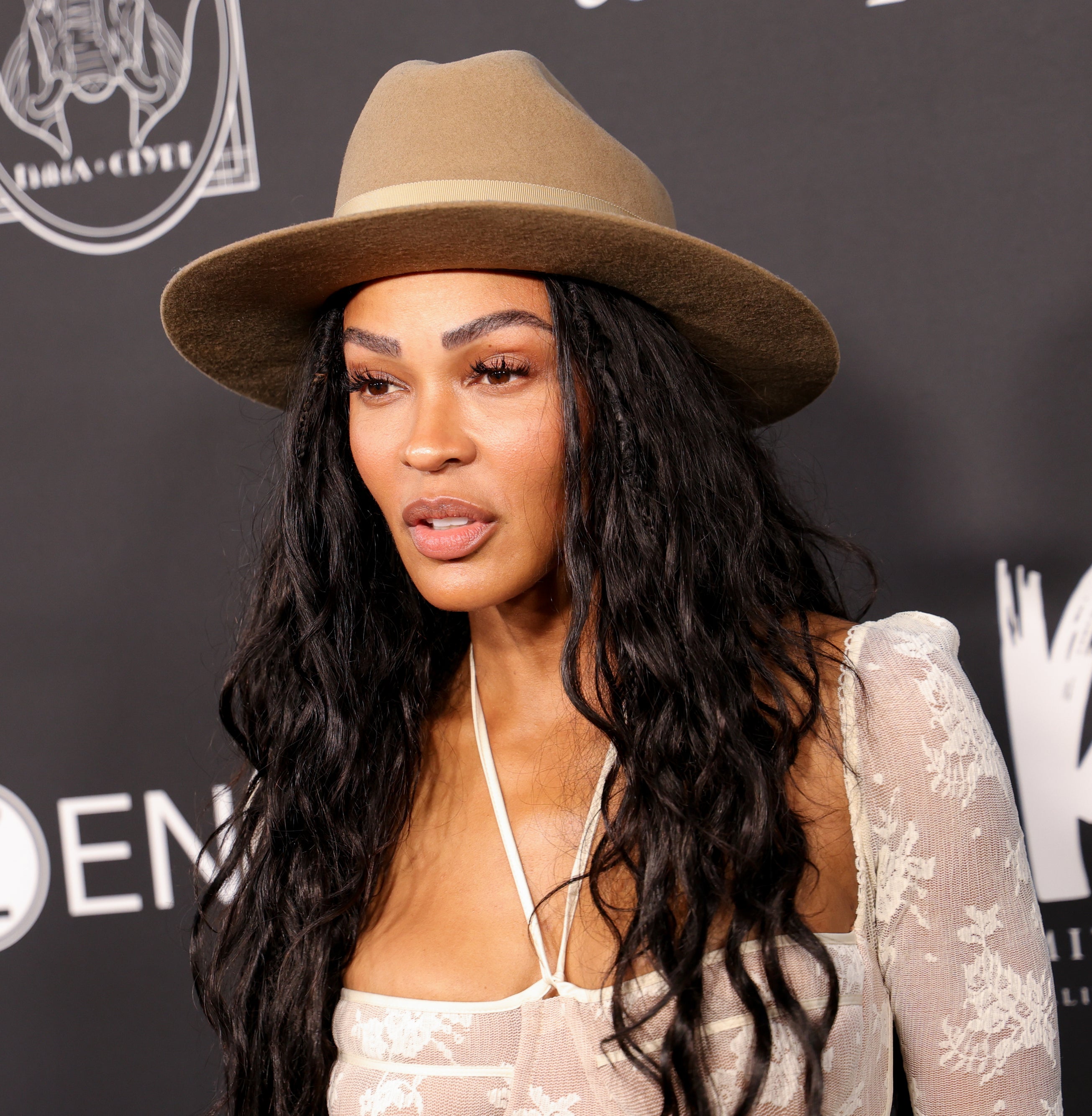 Meagan Good poses on the red carpet, wearing a beige wide-brimmed hat and a white lace top, at a public event backdrop with various logos