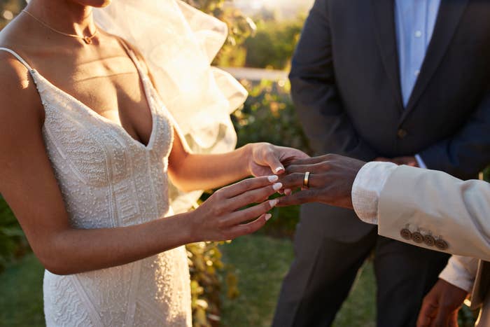 A bride in an intricate lace gown places a wedding ring on the groom's finger during an outdoor ceremony, with an officiant in a suit looking on