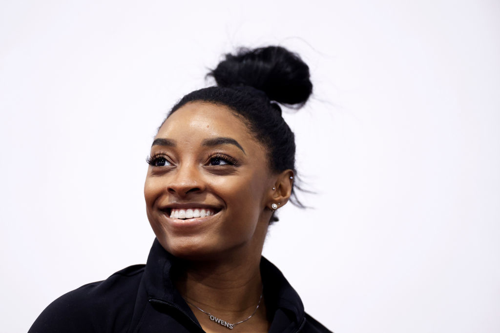 Simone Biles smiles, wearing a black jacket and a necklace with a visible inscription, hair in a bun