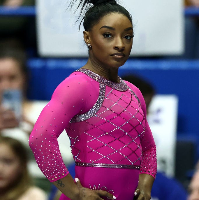 Simone Biles stands confidently in a sequined leotard at a gymnastics event. She wears her hair styled in a high bun