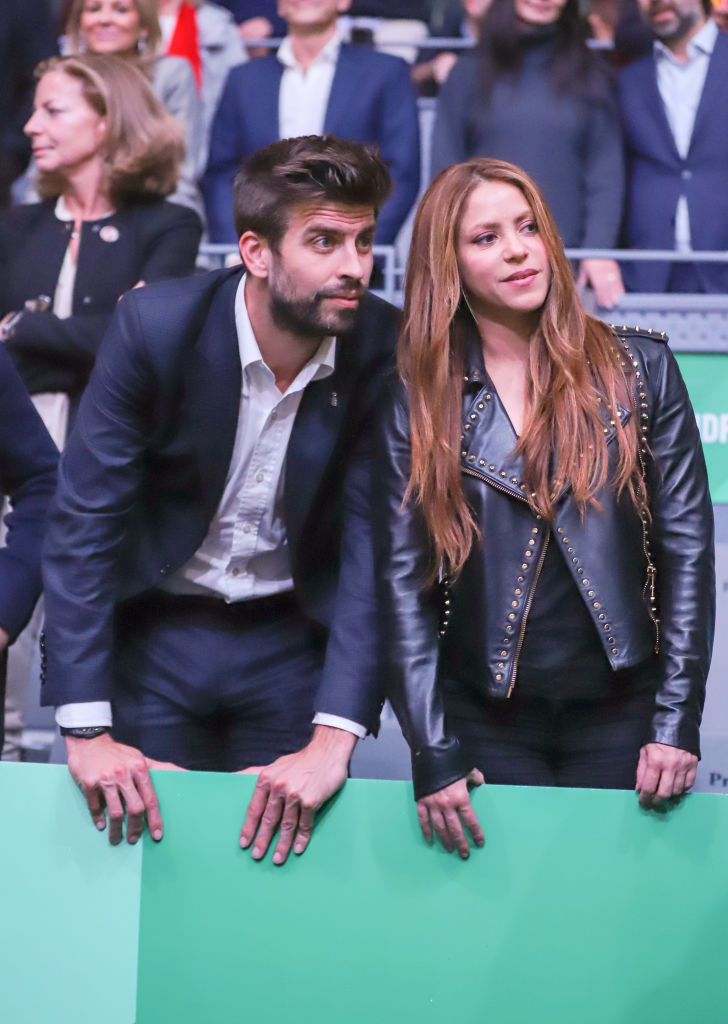 Gerard Piqué and Shakira leaning over a barrier at an event. Piqué is in a suit, and Shakira is wearing a studded leather jacket